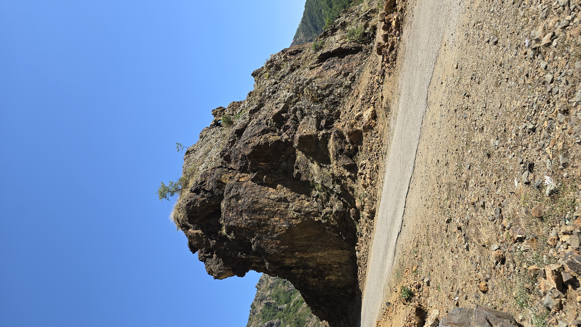 A winding paved road curves around a large, imposing rock formation in Madzharovo, Bulgaria. The rock is rugged and appears to be composed of reddish-brown and darker brown rock with patches of green vegetation and dry grass growing on its surface. Two small trees are visible on the crest of the rock formation. The road is bordered by dry, rocky terrain with scattered stones and sparse dry grass. In the background, hills covered with green vegetation rise under a clear blue sky. The overall impression is a natural, arid landscape with a man-made road cutting through it.