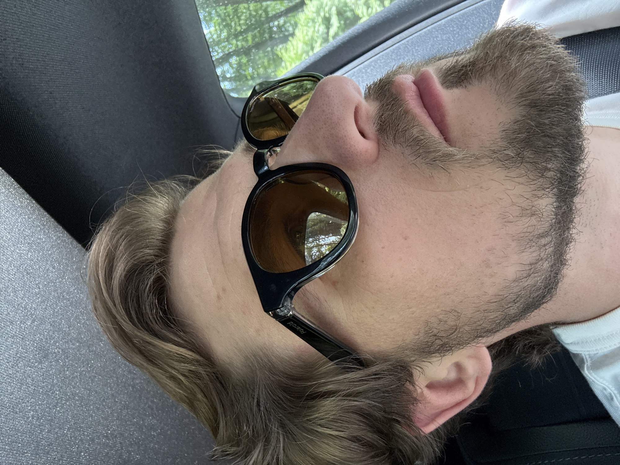 The image shows a close-up, side profile of a person wearing sunglasses and a white t-shirt, likely inside a vehicle. The person has light brown hair, a light beard, and is looking to the right. The sunglasses have black frames and amber lenses, reflecting trees and sky. The interior of the vehicle, including the headliner and part of a seat, is visible. The scene is outdoors, with greenery and trees visible through the vehicle window. The location is identified as Krubki-Górki, Poland.