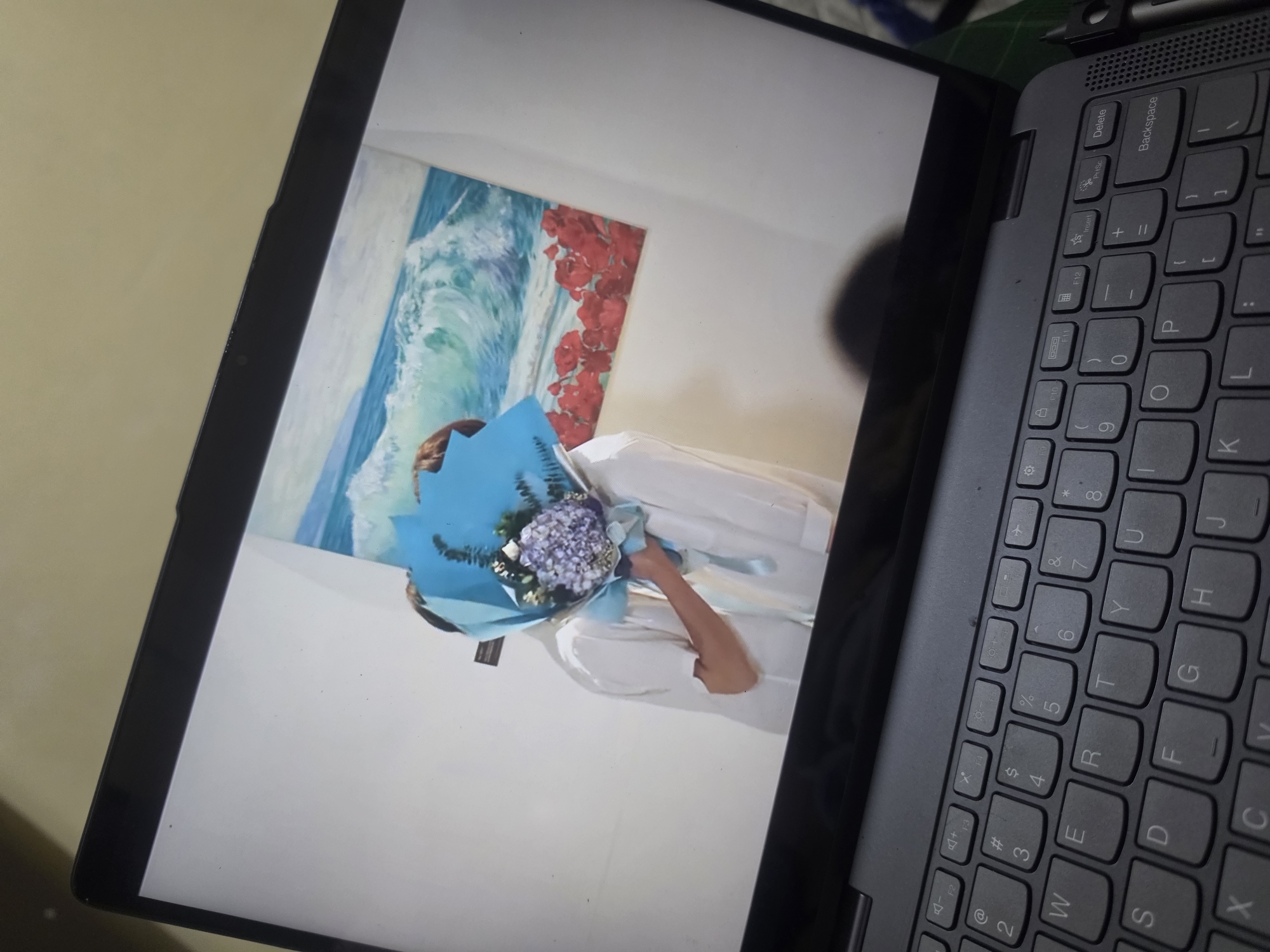 The image depicts a laptop screen displaying a photograph. The photograph shows a person holding a bouquet of flowers wrapped in blue paper. In the background of the photograph is a wall with a painting of ocean waves and red flowers. The laptop's keyboard is visible in the foreground. The scene appears to be indoors. The image was taken in Pintomakmur, Indonesia.
