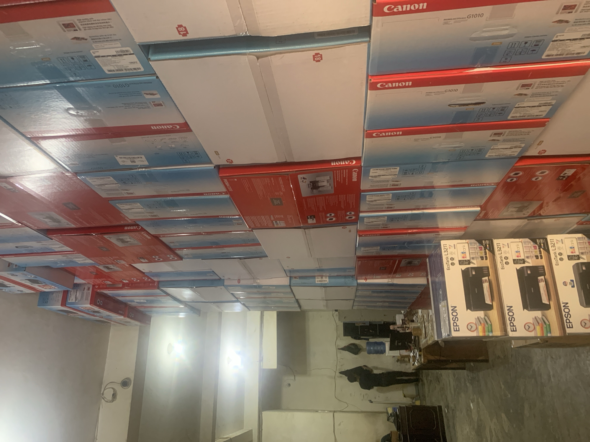 The image depicts a storage room filled with stacked boxes, likely belonging to an electronics store or distributor, in Palembang, Indonesia. The room is dimly lit, with overhead fluorescent lights providing illumination. The walls are painted a light color, and the floor appears to be concrete.
The dominant subjects in the image are numerous cardboard boxes, primarily displaying branding for "Canon" and "Epson." These boxes are stacked high, forming a significant portion of the visual field and suggesting a large inventory. Many of the boxes appear to be for printers or related office equipment. The "Canon" boxes are predominantly red and white or blue and white, while the "Epson" boxes are blue and white and feature images of printers and ink bottles. Some boxes show product names such as "PIXMA G1010" and "EcoTank L3211."
In the left portion of the image, a single individual, appearing to be a male, is visible. He is wearing dark clothing and is standing facing away from the camera, seemingly engaged in some activity within the room. He is positioned near a desk laden with various items, including a computer monitor, papers, and a water dispenser. The desk area suggests a workspace or administrative section within the storage facility.
The overall impression is one of an organized, albeit somewhat cluttered, storage environment for electronic goods. The presence of numerous stacked boxes and a single worker indicates a commercial or logistical operation.