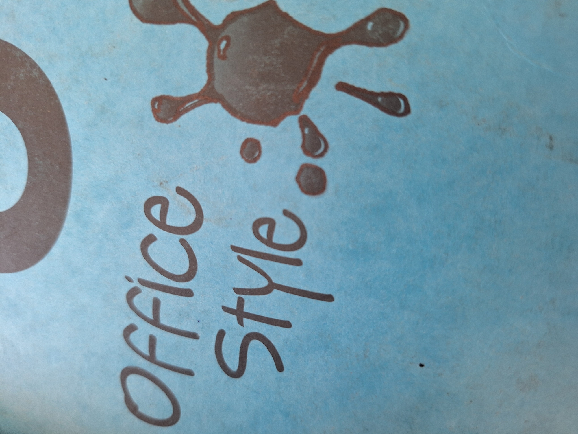 The image is a close-up of a light blue textured surface with dark brown text and ink splatters. The text reads "Office Style" in a casual, handwritten font. To the right of the text, there is an artistic representation of ink splatters, appearing to be a stylized splash or drop with smaller accompanying drops. The background is a uniform light blue. There is no discernible location in Varna, Bulgaria, or any people or actions present in the image. The image is a still shot of graphic elements on a surface.