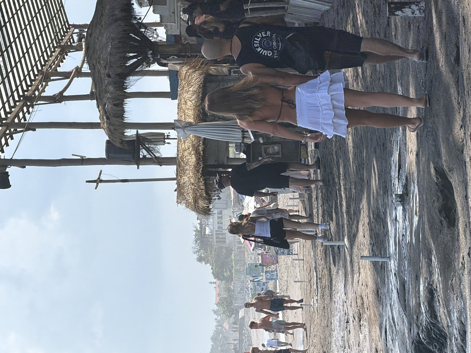 The image depicts a sandy beach scene in North Kuta, Indonesia. In the foreground, a couple walks away from the camera. The woman is wearing a white bikini top and a white ruffled skirt, while the man wears a black tank top with "Quiksilver" printed on it and black shorts. They are walking on a dark, wet, rocky surface with some standing water.
Further in the background, a crowded beach is visible with numerous people, many of whom appear to be walking along the shoreline. Various beachside structures, including huts with thatched roofs and wooden frameworks, are present. Umbrellas and signs are scattered across the sand. The sky is bright and hazy, suggesting it is daytime. The overall atmosphere appears to be one of a bustling tourist beach.