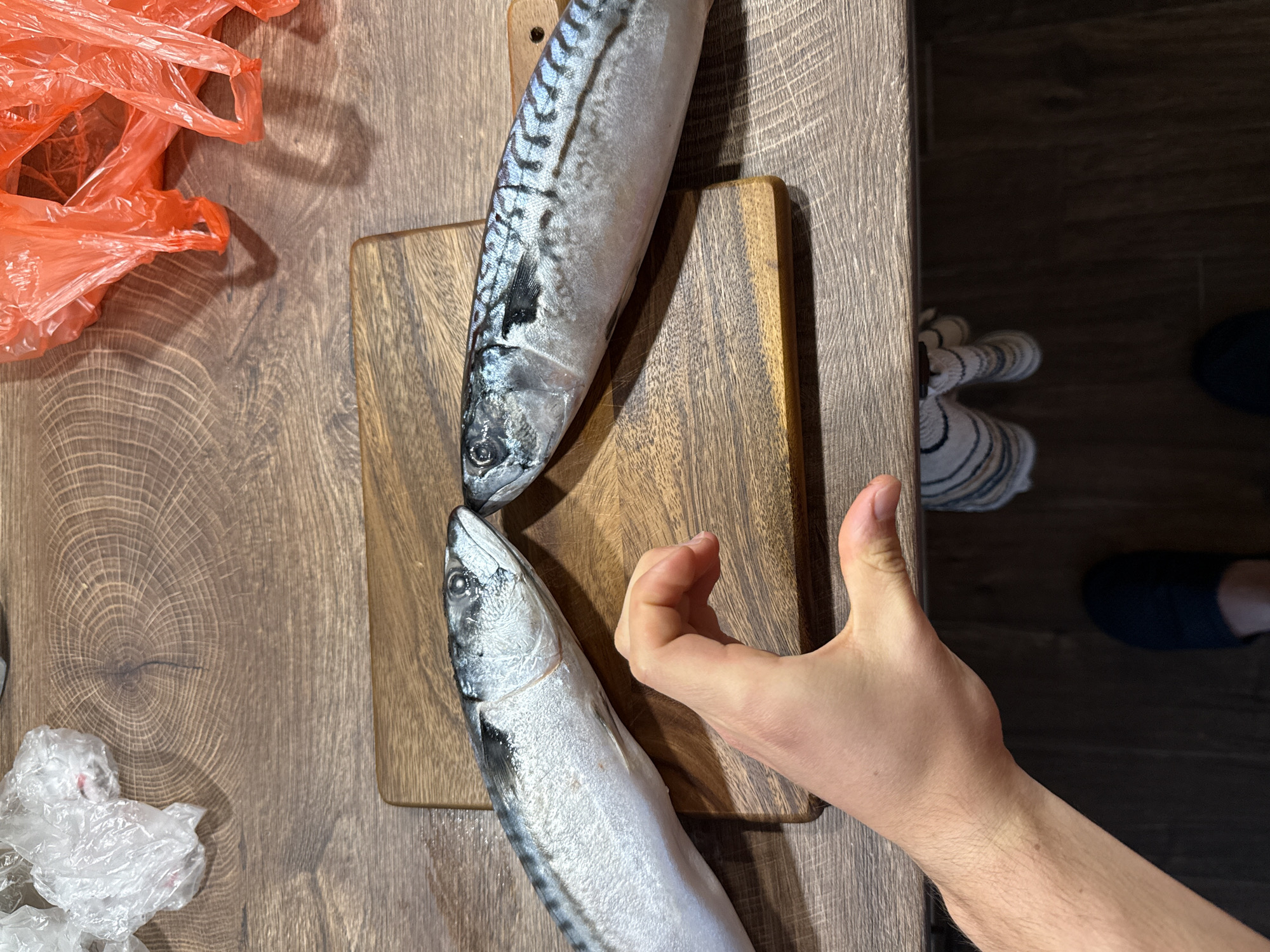 A top-down, slightly angled shot captures an indoor scene, likely a kitchen or food preparation area. The main subjects are two whole, raw mackerel fish arranged on a dark wooden cutting board, which rests on a lighter brown wooden table or countertop featuring a distinct wood grain.
The first mackerel lies horizontally across the upper part of the cutting board, its head facing left. The second fish is positioned diagonally below it, with its head pointing towards the bottom-left, almost touching the tail of the first fish. Both fish exhibit their natural silver-grey coloration with characteristic dark, wavy stripes along their backs.
A fair-skinned hand extends into the frame from the right side, giving a clear "thumbs up" gesture, implying satisfaction or approval, possibly regarding the fish. The forearm of the hand is also visible.
In the upper left portion of the image, several crinkled, translucent red plastic bags are clustered together, casting shadows on the wooden surface. Below these, in the bottom-left corner, a smaller pile of crinkled white plastic bags is visible. To the bottom right, a glimpse of a striped cloth or towel in white and blue tones, and a dark blue shoe or slipper on a darker floor, can be seen beyond the edge of the table. The overall lighting suggests an indoor setting with a visible light source creating shadows. This scene is located in Sofia, Bulgaria.