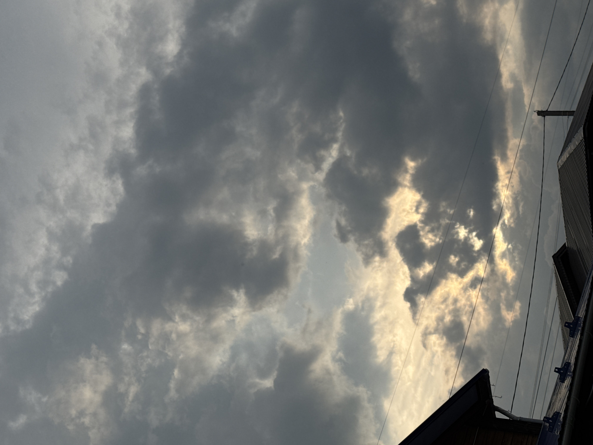 The image captures a dramatic sky filled with dark, textured clouds, with patches of bright sunlight breaking through. The clouds are a mix of deep grays and lighter, illuminated areas, suggesting dynamic weather conditions, possibly approaching rain or a storm.
In the lower right portion of the frame, the roofline of a building is visible, constructed with corrugated metal siding. A dark, sloped roof with some framing is also present in the foreground. Utility wires, strung between poles, crisscross the sky, adding a man-made element to the natural scene. One utility pole is clearly visible.
The time of day appears to be late afternoon or early evening, given the angle of the sunlight and the dramatic cloud formations. The overall mood is somewhat somber and intense, dominated by the powerful presence of the sky. The setting appears to be a rural or suburban area, indicated by the building structures and overhead wires, consistent with a location in Старое Михайловское, Russia. There is no visible text in the image.