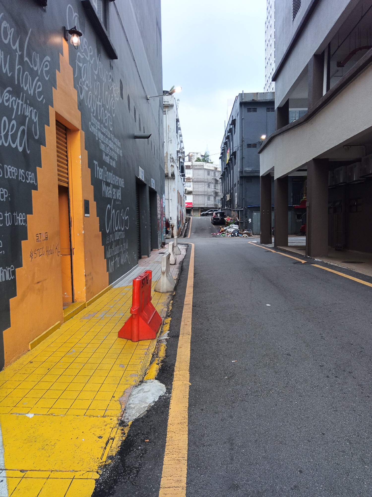 This is a medium-angle, eye-level, full shot of a narrow street in Kuala Lumpur, Malaysia. The street is flanked by buildings on both sides. On the left, a building with a dark gray facade features white chalk lettering and a bright yellow, tiled sidewalk. A bright red traffic barrier is positioned on the sidewalk. On the right, a modern building with exposed concrete columns and balconies lines the street. The asphalt road is marked with yellow lines. In the distance, a pile of refuse is visible near a black car parked on the left side of the road. The sky is overcast.