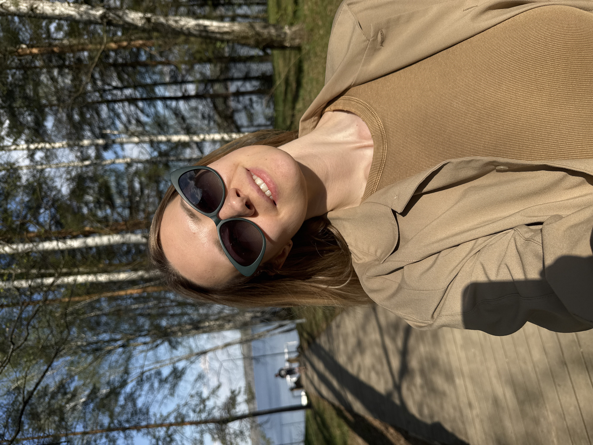 A woman is posing outdoors in a park-like setting in Chaikovsky, Russia. She's wearing sunglasses, a light-brown ribbed top, and a tan jacket. The background features trees with white bark, possibly birch trees, against a blue sky. A body of water is visible in the distance with people visible on a dock. The woman is smiling, and the image is well-lit, suggesting it was taken during the day. The scene conveys a relaxed, outdoor environment.