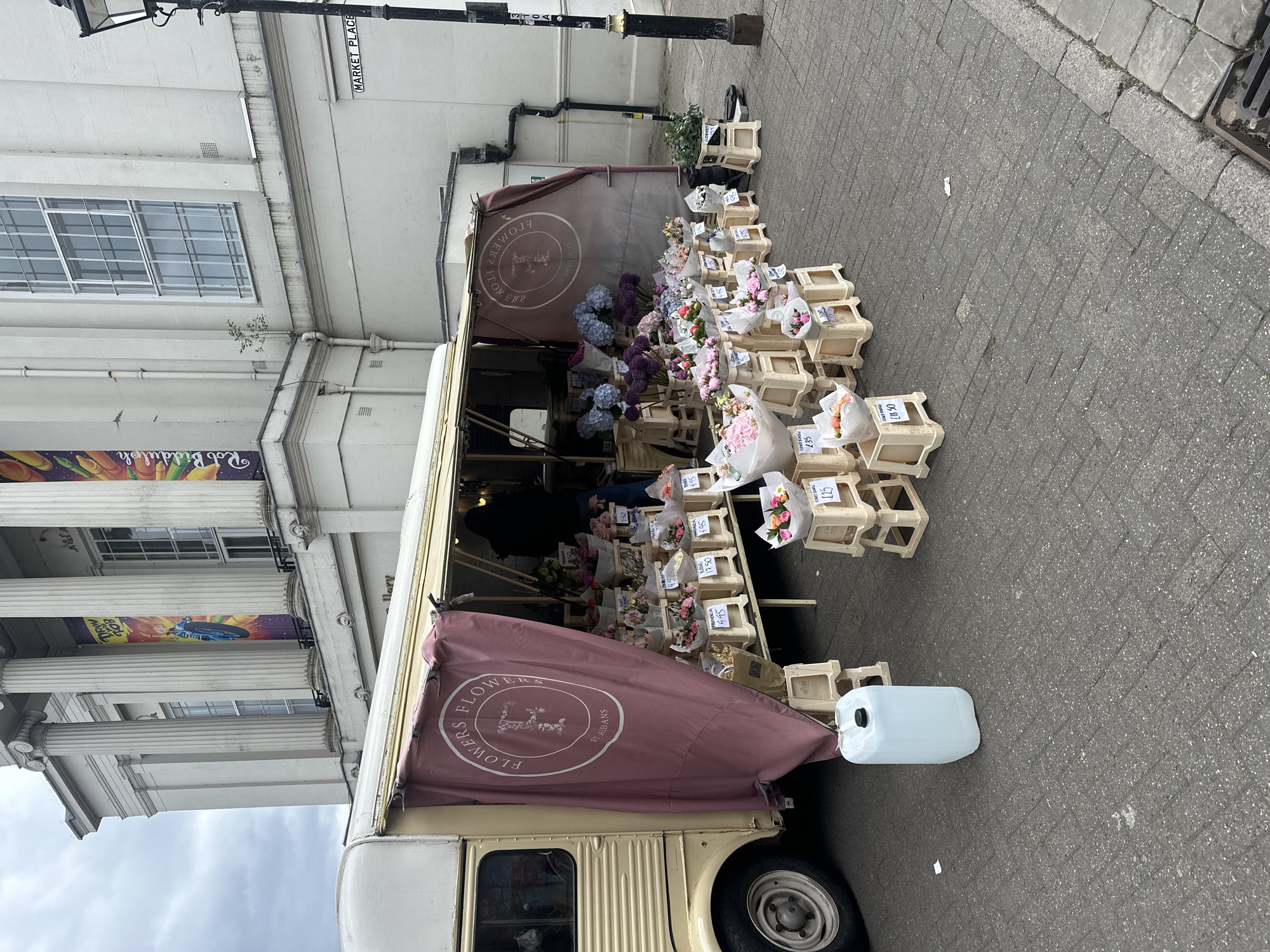 The image shows a flower stall situated next to a building with classical architectural elements, including columns and large windows. A sign reading "MARKET PLACE" is visible on the building. The stall is a van-like structure with a canopy displaying the text "FLOWERS FLOWERS ST. ALBANS" in a circular logo. Numerous bouquets of flowers are arranged in wooden crates, with price tags displayed on some of them. A white plastic container is positioned beside the stall. The scene is shot at street level on a paved area.