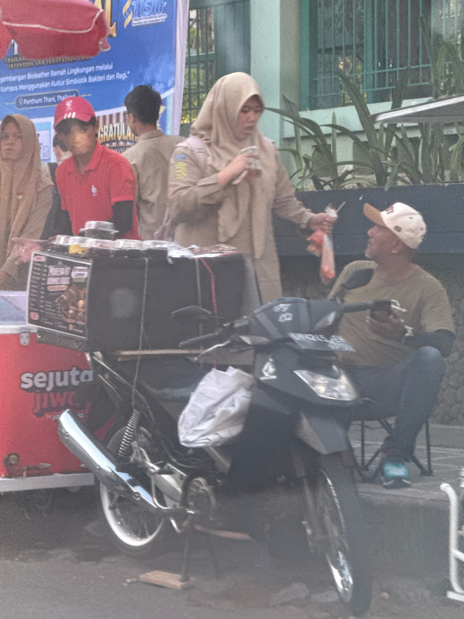 In the city of Surabaya, Indonesia, a street scene unfolds with several individuals and objects arranged in a casual outdoor setting. In the foreground, a motorcycle with a large black box mounted on its rear serves as a mobile food cart. The box has a sign with text that is partially visible, including "MORENA-87" and what appears to be a menu. A red cooler with white text reading "sejuta jiwe" is positioned to the left of the motorcycle.
Two individuals are interacting near the food cart. A woman wearing a beige hijab and a beige jacket is holding a clear plastic cup with a straw and appears to be drinking. To her right, a man wearing a light-colored baseball cap and a green t-shirt is seated, looking at his phone. He is wearing blue jeans. Another person, wearing a red collared shirt and a red cap, is partially visible behind the motorcycle. Further to the left, a woman with a beige hijab is also partially visible. In the background, a banner with text in Indonesian and English is displayed, and behind that, a building with windows and greenery is seen. The overall environment suggests a street or market area, possibly during the daytime.