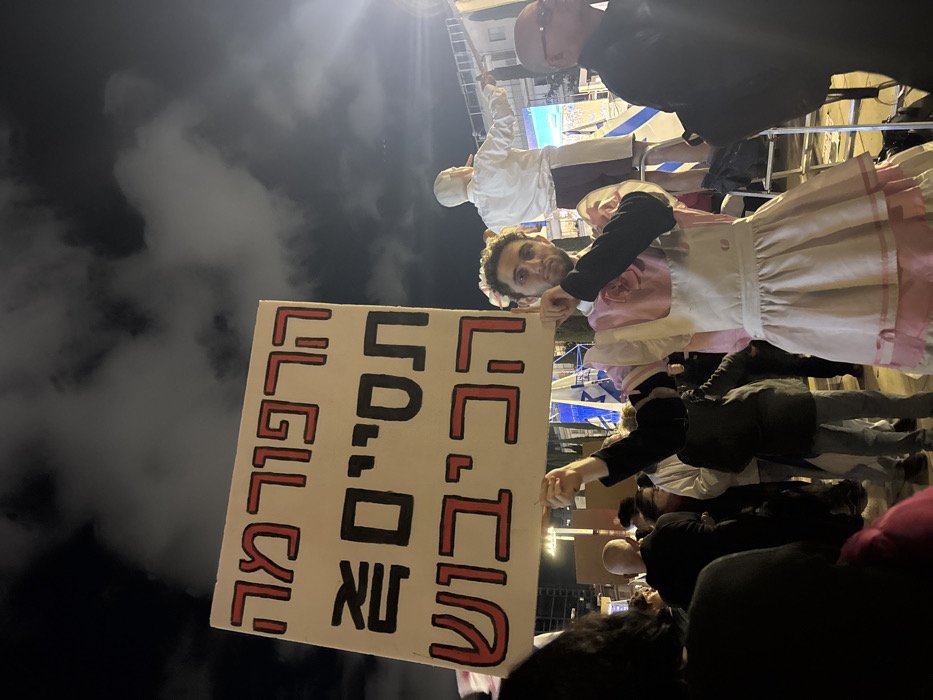 A person in a pink and white maid outfit holds a sign with Hebrew text. The sign has red outlines and black text. Other individuals surround the subject, and additional figures are standing on an elevated platform behind them. The sky in the background is dark with some cloud cover. The photo is taken at night, and the lighting is somewhat dim.