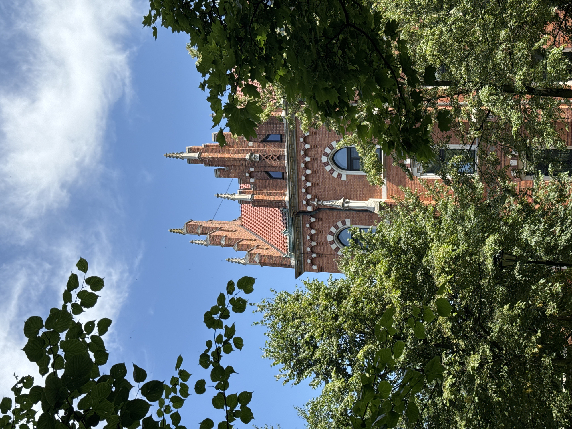 This is a medium-angle, eye-level shot of the exterior of Jagiellonian University's Collegium Minus building in Krakow, Poland, viewed through the foliage of trees. The building, constructed of red brick, features Gothic Revival architecture with decorative stone trim, pointed arch windows, and turrets topped with finials. The sky above is a bright blue with scattered white clouds. Lush green trees with dense foliage frame the building, obscuring parts of its facade and lower levels. A black lamppost is visible amongst the trees in the lower right quadrant of the image. The scene depicts a sunny day in an urban environment, focusing on architectural detail contrasted with natural elements.