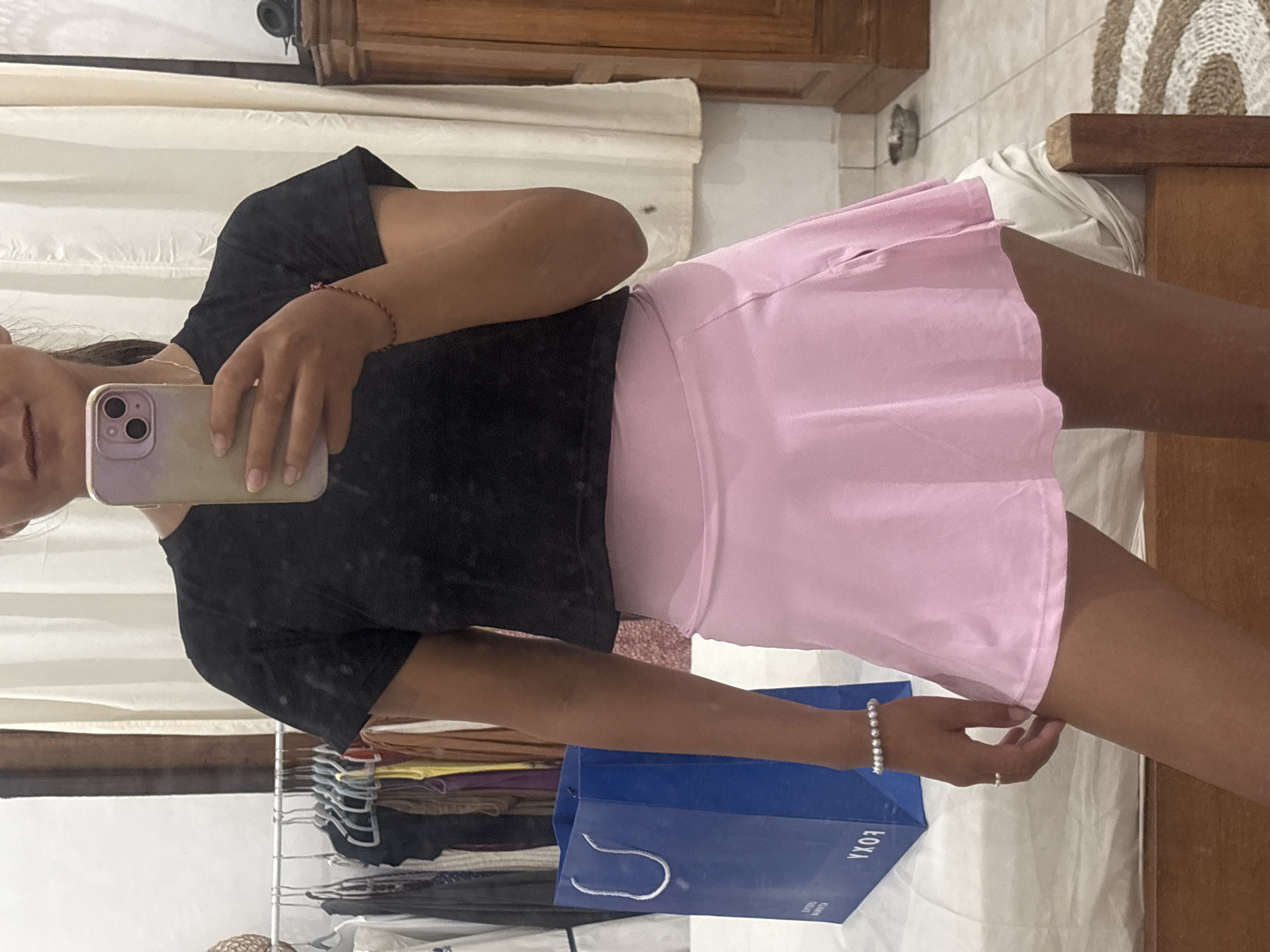 A person stands indoors, holding a phone and looking at the camera's screen, with a rack of clothes and a blue shopping bag nearby. The location appears to be Kuta Selatan, Indonesia.