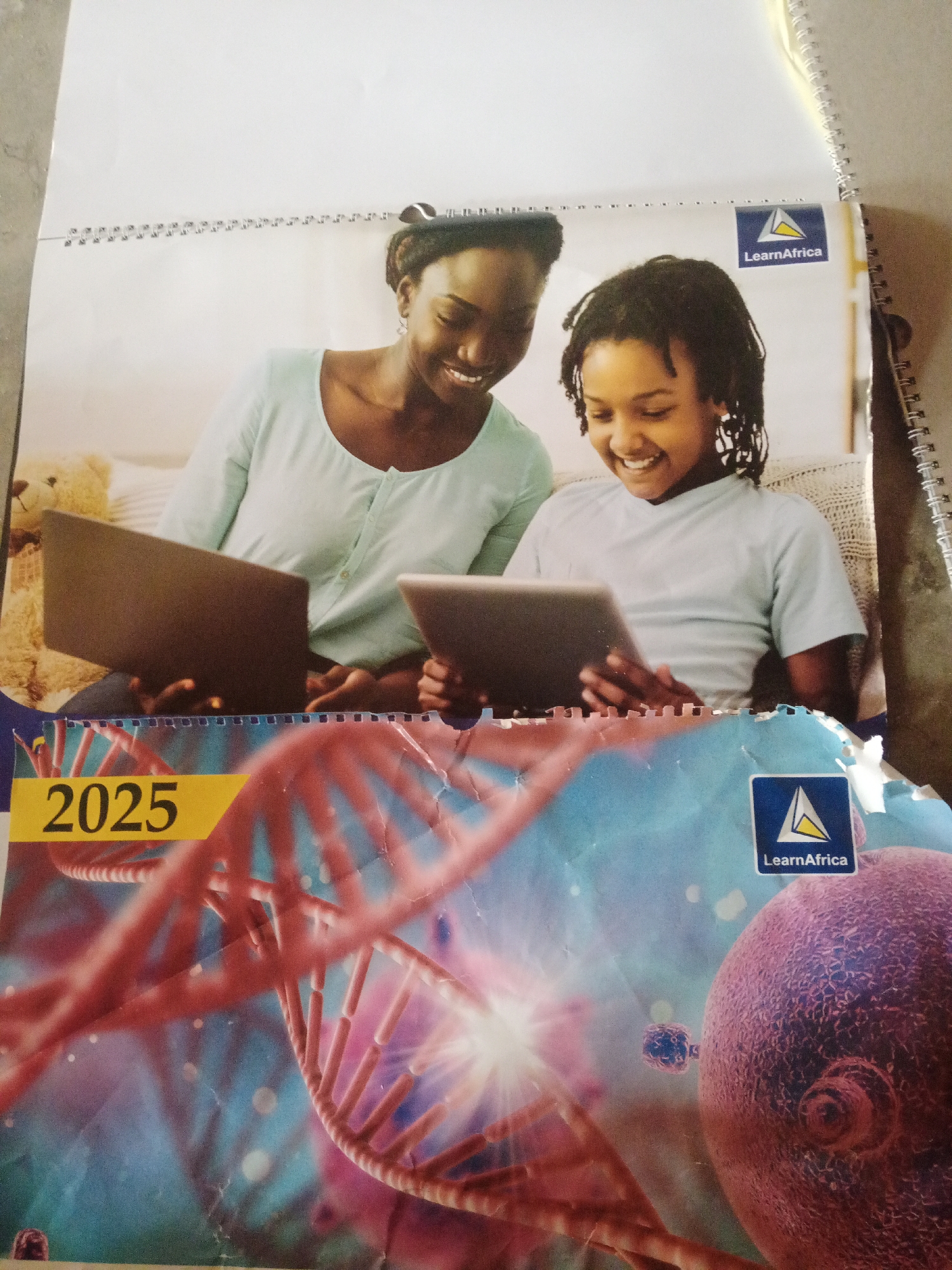 The image is a calendar or poster featuring two individuals, likely a mother and daughter, looking at electronic devices. The top portion shows them sitting and smiling while interacting with a laptop and a tablet. A teddy bear is visible to the left of the woman. In the upper right corner, there is a logo with the text "LearnAfrica". The bottom portion of the image displays a graphic of a DNA helix and a cell, with the year "2025" prominently featured in yellow. A second "LearnAfrica" logo is present in the lower right corner. The overall aesthetic suggests an educational or promotional context, possibly related to science or technology for students. There is no indication of the specific location within Jalingo, Nigeria.