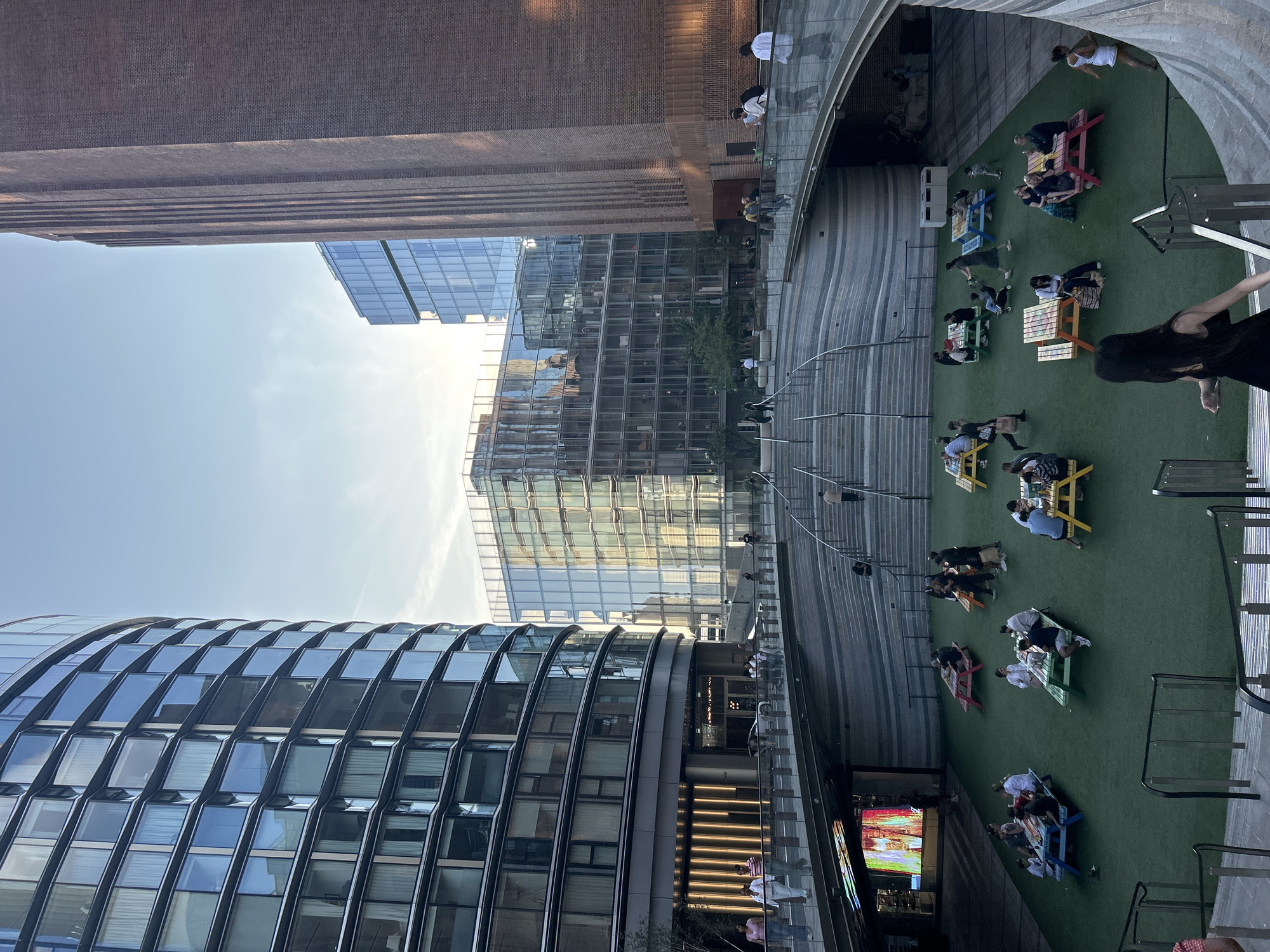 The image captures a view of the Tate Modern's outdoor space in London, United Kingdom. Several contemporary buildings with glass facades flank a brick building, with a large staircase structure in the foreground. People are seated at picnic tables scattered across a bright green, artificial turf area. The sky is clear with some wispy clouds, and the lighting suggests it is late afternoon or early evening.