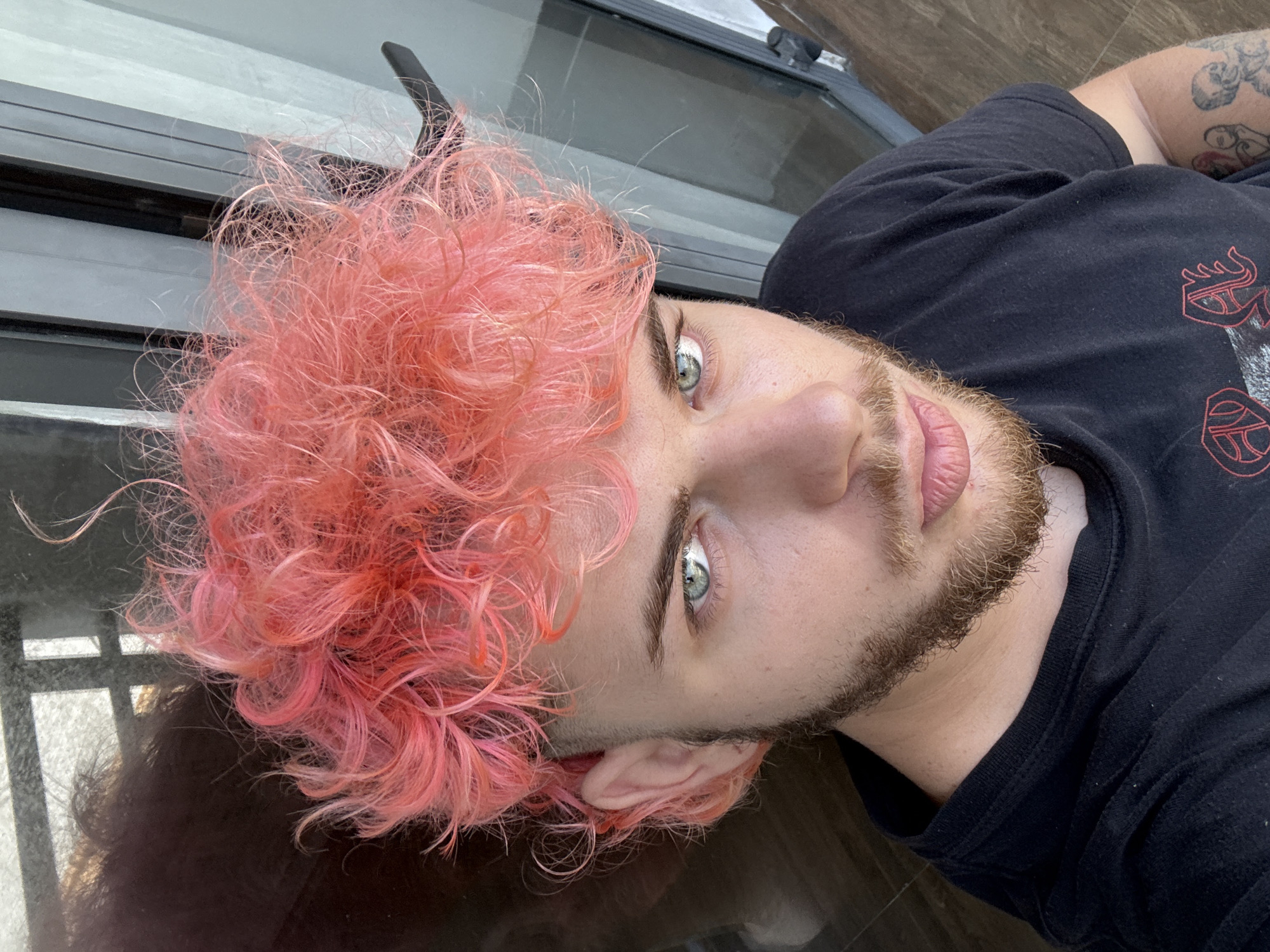 A close-up, eye-level shot depicts a fair-skinned individual with light-colored eyes and a beard. The person has dyed, bright pink, curly hair. They are wearing a black t-shirt with a red and white graphic design. A partial view of a tattoo is visible on their arm. The background appears to be a reflective surface, possibly a table or window, showing reflections of a window frame. The location, Đà Nẵng, Vietnam, cannot be determined from the image alone.
