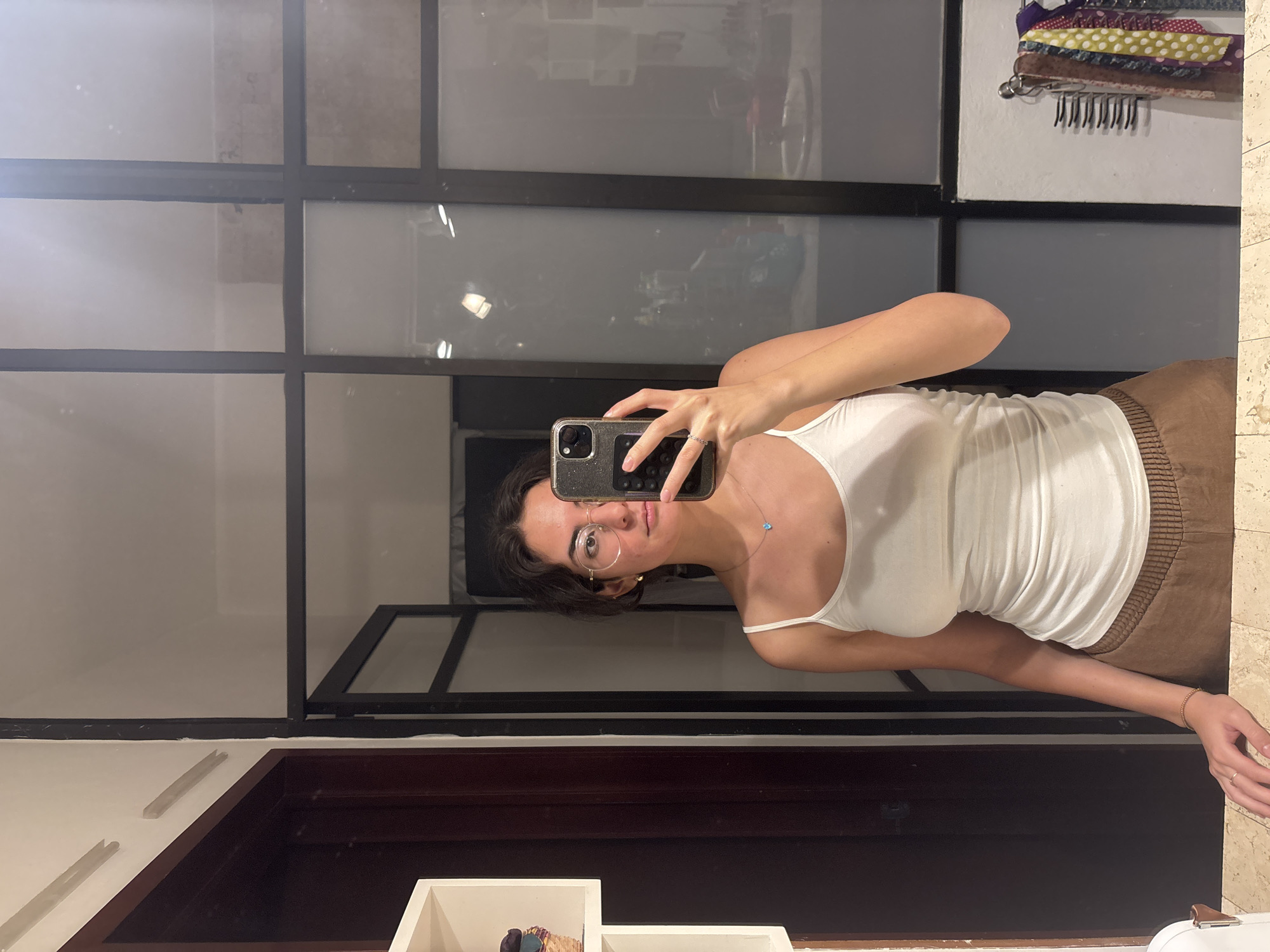 A woman is taking a selfie in a bathroom or dressing area with a large mirror. She is wearing a white tank top and brown bottoms. She has round glasses and a necklace. The mirror reflects a multi-pane window with a view of what seems to be a city street, potentially showing a bicycle. There's a hanging organizer with scarves or fabric pieces on the right side of the mirror. In the foreground, below the mirror, is a dark wooden surface, and some white shelving or cabinetry is visible. The location might be Puebla, Mexico.