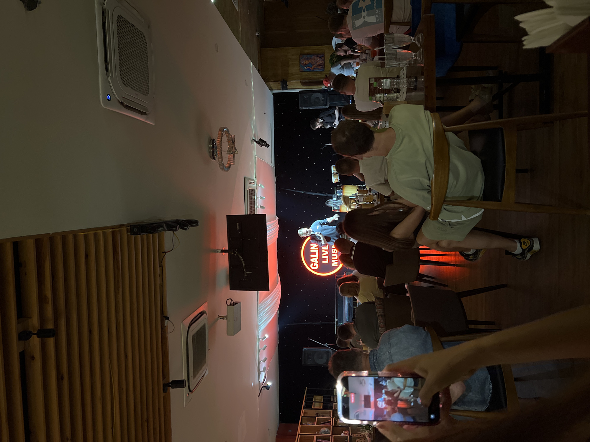 The image captures a live music performance at a venue, likely a bar or restaurant in Nha Trang Ward, Vietnam. Patrons are seated and observing the show. One person is using a smartphone to record the performance. The band is visible on a stage with a neon sign reading "GALIN LIVE MUSIC." Speakers and lighting fixtures are evident. The interior includes air conditioning units, decorative lighting, and wooden paneling. Patrons are seated at tables and engaging with each other or observing the show.