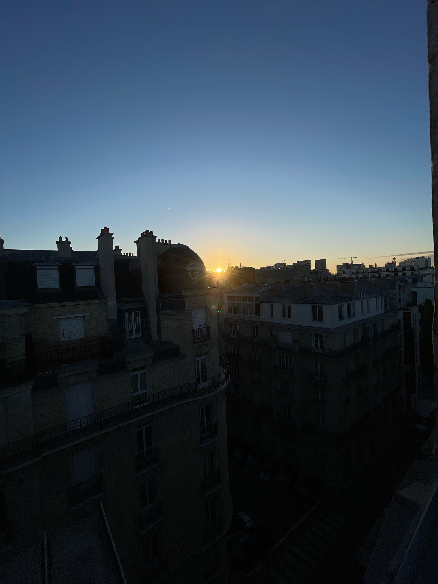 An aerial view captures the Parisian skyline at sunrise. The sun, partially obscured by buildings, casts a warm glow across the scene. Residential buildings, characterized by their mansard roofs and chimneys, dominate the foreground. In the distance, taller structures form the silhouette of the city. The sky transitions from a deep blue at the top to a lighter, warmer hue near the horizon.