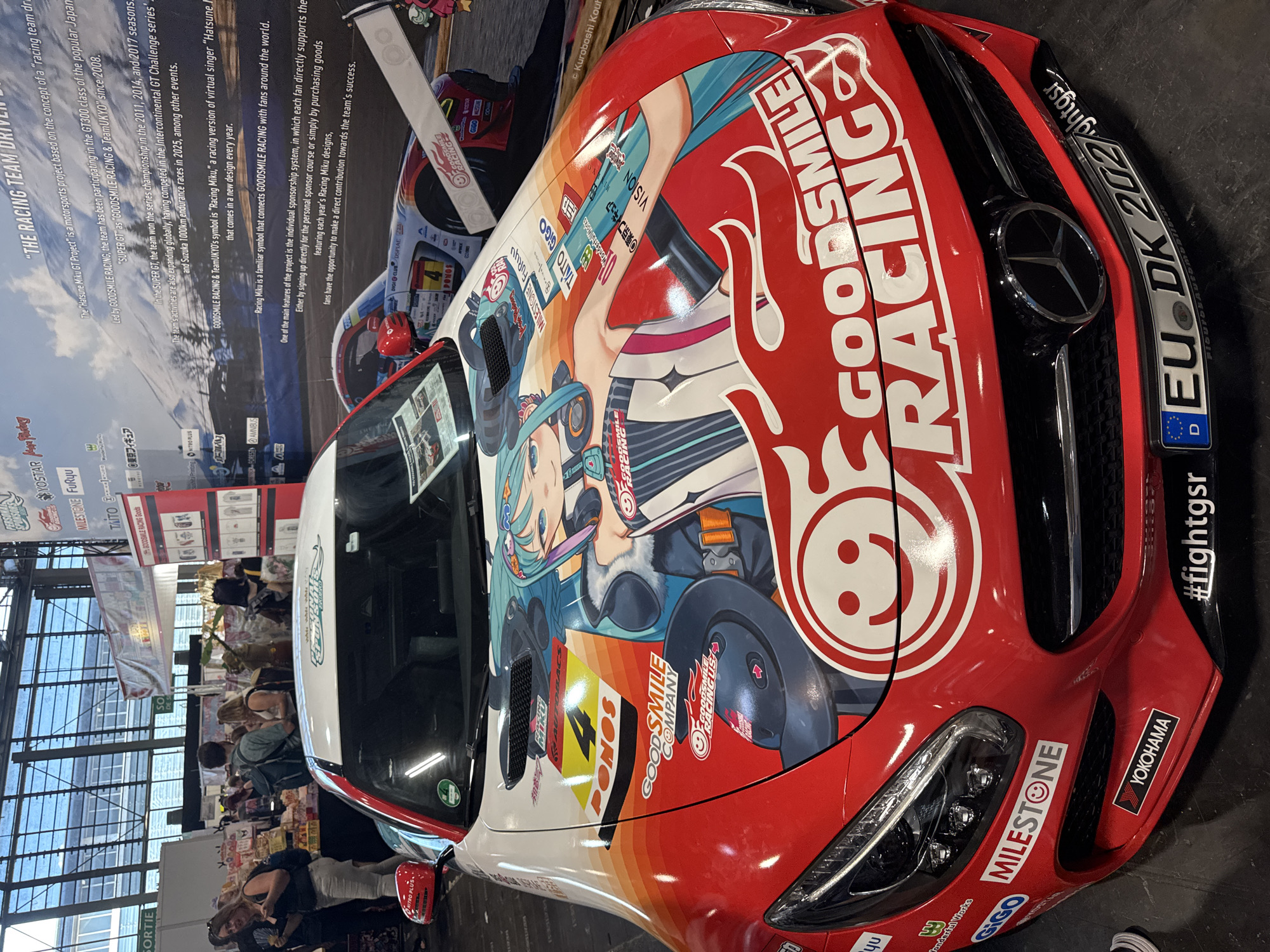 The image shows a red Mercedes-Benz car with a large anime-style graphic on its hood, parked indoors. The car is heavily branded with "GOODSMILE RACING" text and logos. In the background, there are booths and displays associated with a convention or exhibition, suggesting a public event. People are visible in the background, browsing the exhibits. The context provided indicates this scene is from the Paris Manga & Sci-Fi Show at the Paris-Nord Villepinte Exhibition Centre in Villepinte, France. The car itself is the primary subject, adorned with vibrant graphics of an anime character, likely Hatsune Miku, a virtual singer often associated with racing teams. The car's headlights, grille, and license plate are clearly visible. The environment appears to be an exhibition hall with bright lighting and a diverse crowd.