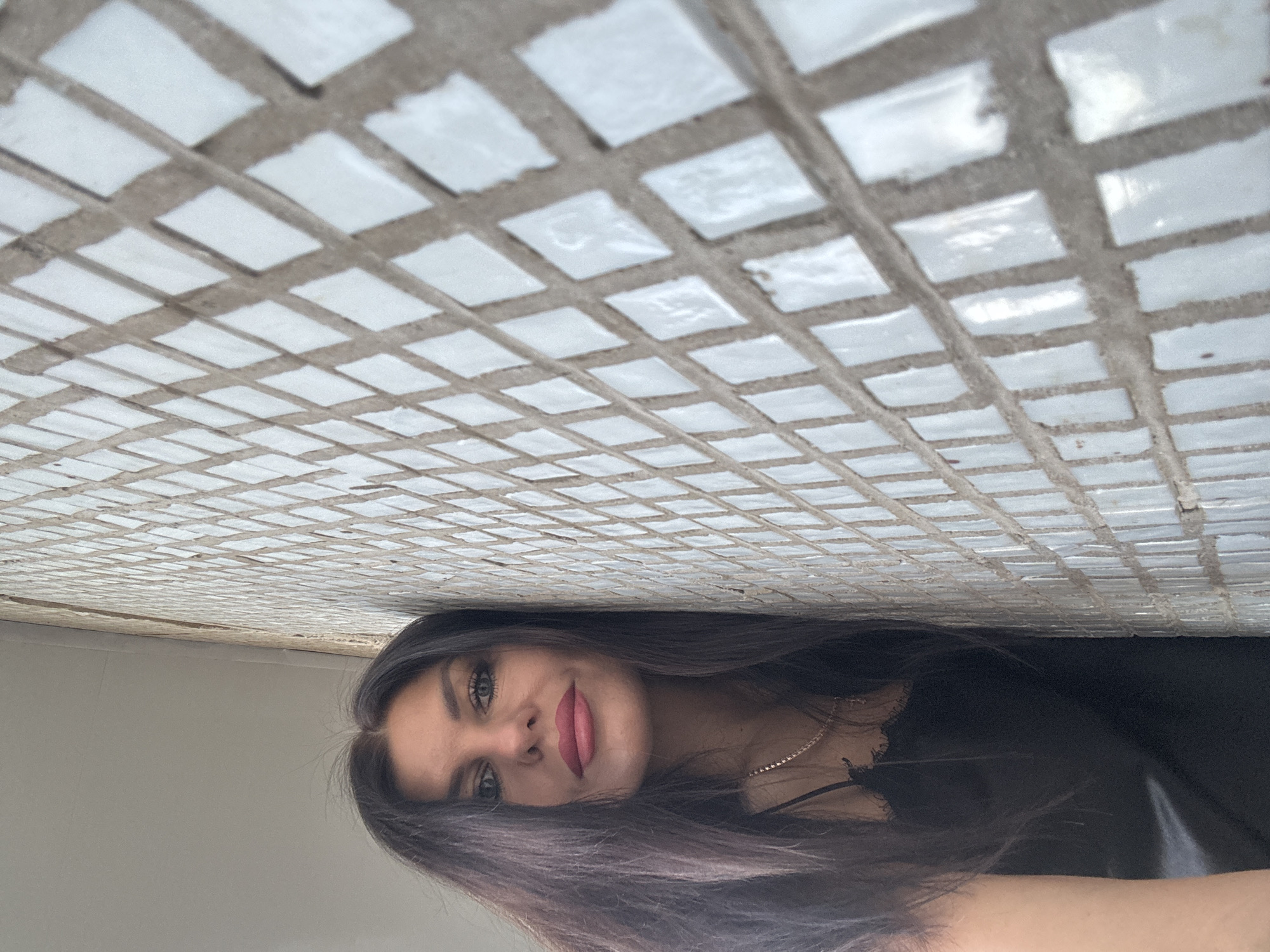 The image displays a portrait of a woman, captured with a vertical orientation where the subject is rotated 90 degrees counter-clockwise.
The primary subject is a young woman with long, dark hair, which has subtle lighter or reddish-purple streaks, framing her face and flowing down her shoulders. She possesses fair skin and features pronounced eyes with visible eyeliner and mascara. Her lips are adorned with bright pink-red lipstick, forming a gentle, pleasant smile as she gazes directly at the camera. She is wearing a dark-colored top, possibly made of satin or silk, with delicate straps and a lace trim visible over her left shoulder and chest. A thin, light-colored necklace is also visible around her neck.
The background features a prominent grid pattern on what appears to be a ceiling or an architectural element directly above her (if the image were properly oriented). This pattern is formed by numerous small, irregularly shaped, light-colored (white/off-white) square tiles separated by dark grey grout lines. Below this tiled section, the background transitions to a plain, smooth surface of a light grey or off-white color.
The lighting is soft and diffused, illuminating the scene evenly without harsh shadows. The time of day appears to be daytime. No text is visible within the image. The general location context is Chaikovsky, Russia.