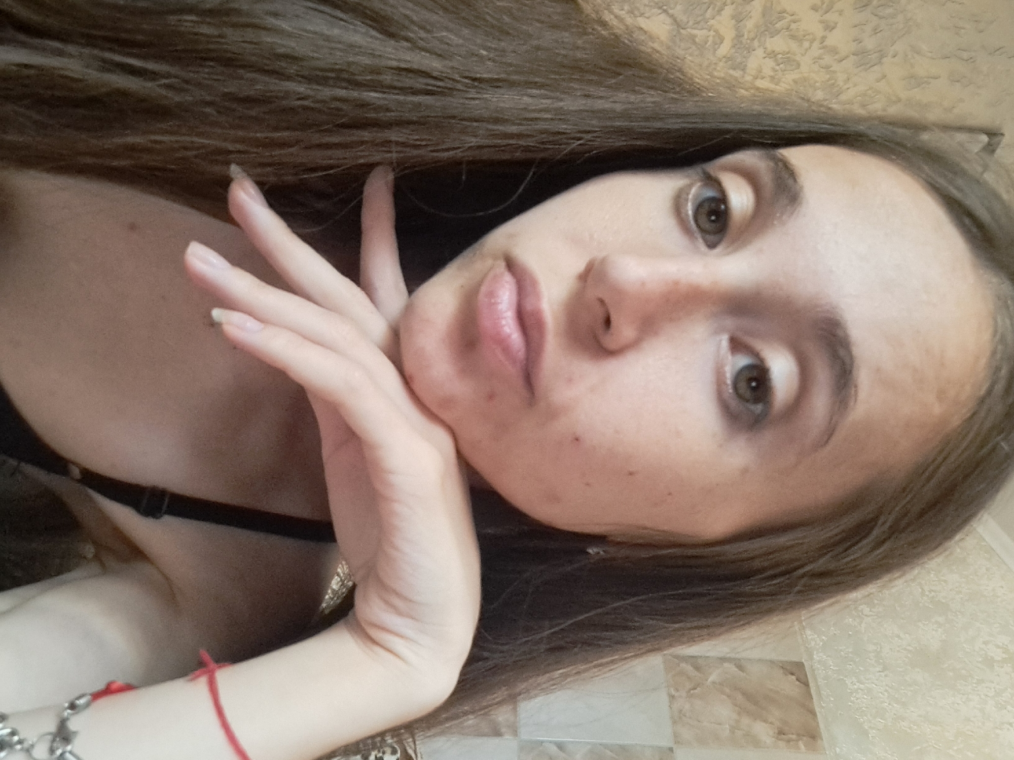 A young woman with long, dark brown hair and light skin is pictured in a close-up shot. She is leaning her head on her hand, with her fingers gently resting on her chin. Her eyes are open and looking towards the camera. She is wearing a black bra strap visible on her left shoulder. A red bracelet is visible on her right wrist, along with a silver charm bracelet. The background appears to be an indoor setting with textured wallpaper and tiled sections. The lighting is moderate, casting soft shadows on her face. The image does not contain any specific information that would allow for its location to be identified within the city of Vypasne, Ukraine.