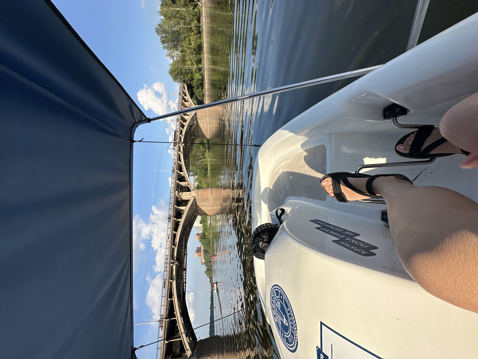 A first-person perspective shot shows a person's legs and feet wearing black sandals, resting on the pedals of a white paddle boat. The boat is on a calm body of water, with a large, arched concrete bridge spanning the river in the background. Trees and buildings are visible on the far bank of the river. The sky is blue with scattered white clouds, and a dark blue canopy of the paddle boat extends over the top of the frame. There are other boats on the water, including a motorboat and a few kayaks, and two individuals are visible on the bridge. The lighting suggests it is daytime, likely late afternoon, given the warm tones reflecting on the water.