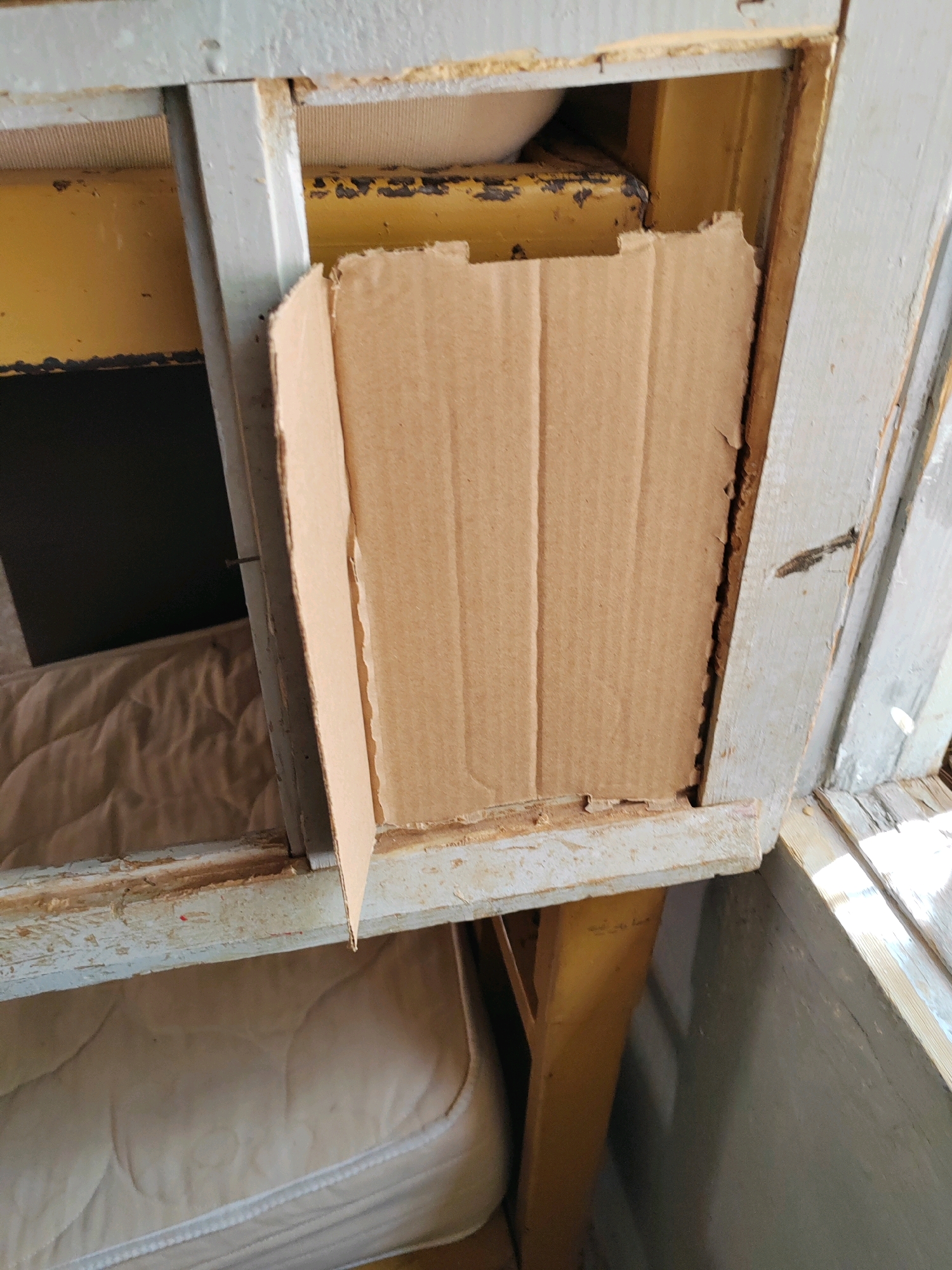 The image shows a close-up of a partially dismantled wooden structure with visible mattresses and a piece of cardboard. The structure appears to be old and weathered, with peeling paint and exposed wood. A piece of brown corrugated cardboard is propped up against the interior of the structure, partially obscuring the view of what lies behind it. The cardboard is roughly cut and has visible fold lines. Beneath the cardboard, the interior of the structure appears to be a shelf or compartment containing what looks like padding or a mattress. Another mattress is visible at the bottom of the frame, suggesting this might be an abandoned or disused living space. The overall scene is one of disrepair and neglect. There are no people or animals visible in the image. The lighting suggests it is daytime, with sunlight casting shadows and highlighting the textures of the materials. There is no information available to determine the specific location within Mougheul, Algeria.