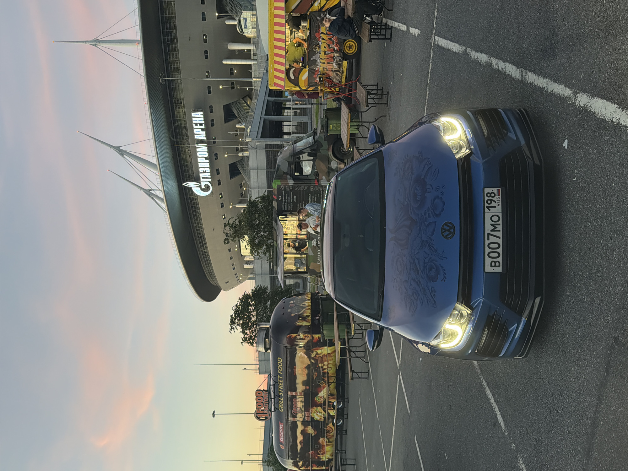 In a parking lot facing the Gazprom Arena in St. Petersburg, Russia, a blue Volkswagen Scirocco is parked centrally, its headlights illuminated. The car's hood features a floral pattern. In the background, several food trucks are set up, with people visible ordering and eating at picnic tables. The sky displays a soft, pastel sunset with pink and blue hues.
