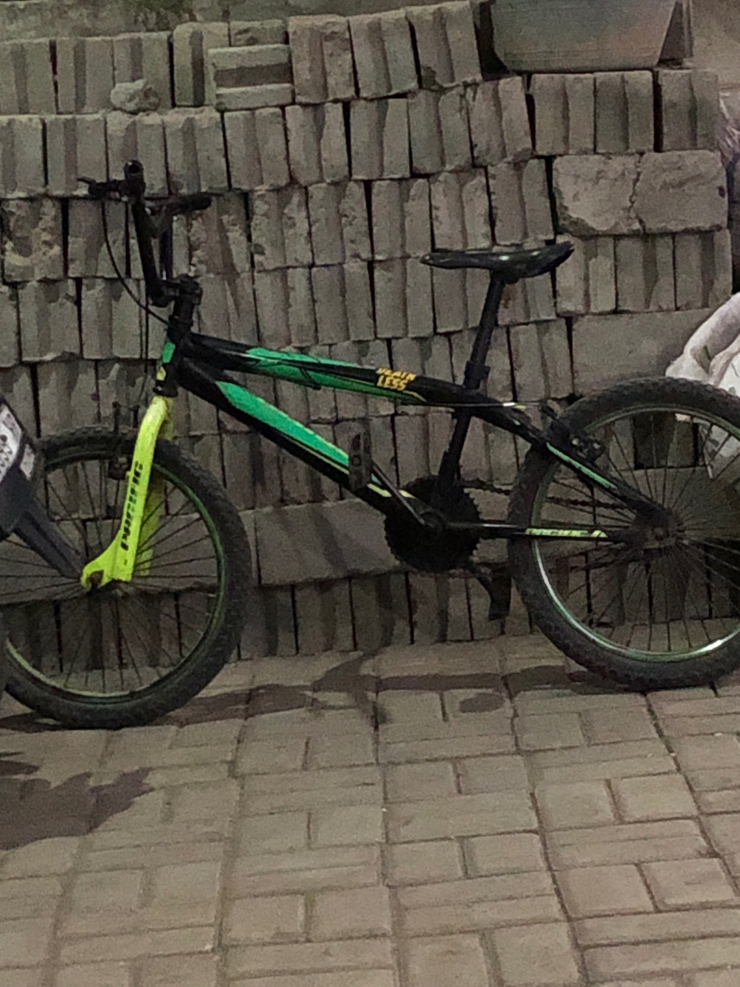 A black and green BMX bicycle is leaning against a wall made of cinder blocks. The bicycle has neon green front forks with "PACIFIC" printed vertically on them. The handlebars are black, and the seat is also black. The bicycle frame is black with green accents and lettering that appears to say "TAIN LESS". The rear wheel is visible, and the bicycle is positioned on a brick-paved ground. The background consists of stacked cinder blocks, some of which are topped with a rounded object. The lighting suggests it may be nighttime or an indoor setting with artificial light. There are no people or animals visible in the image. The location is specified as Keniten, Indonesia.