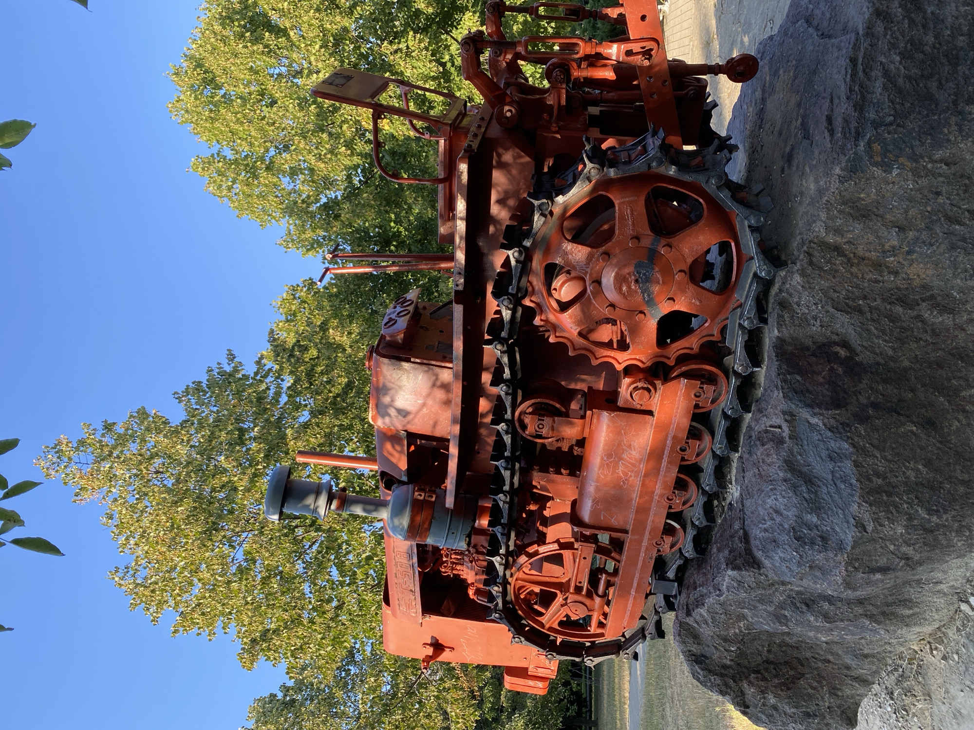 An orange tracked tractor monument is displayed on a large boulder in a park-like setting. The tractor, painted bright orange, features visible tracks, a seat, and an exhaust pipe. The surrounding environment includes green trees with some autumn foliage and a clear blue sky. In the background, there is a paved path and a hint of grass. The scene appears to be outdoors, likely in a public space. The specific location in Chișinău, Moldova, is a park, likely dedicated to agricultural machinery or as a historical monument.