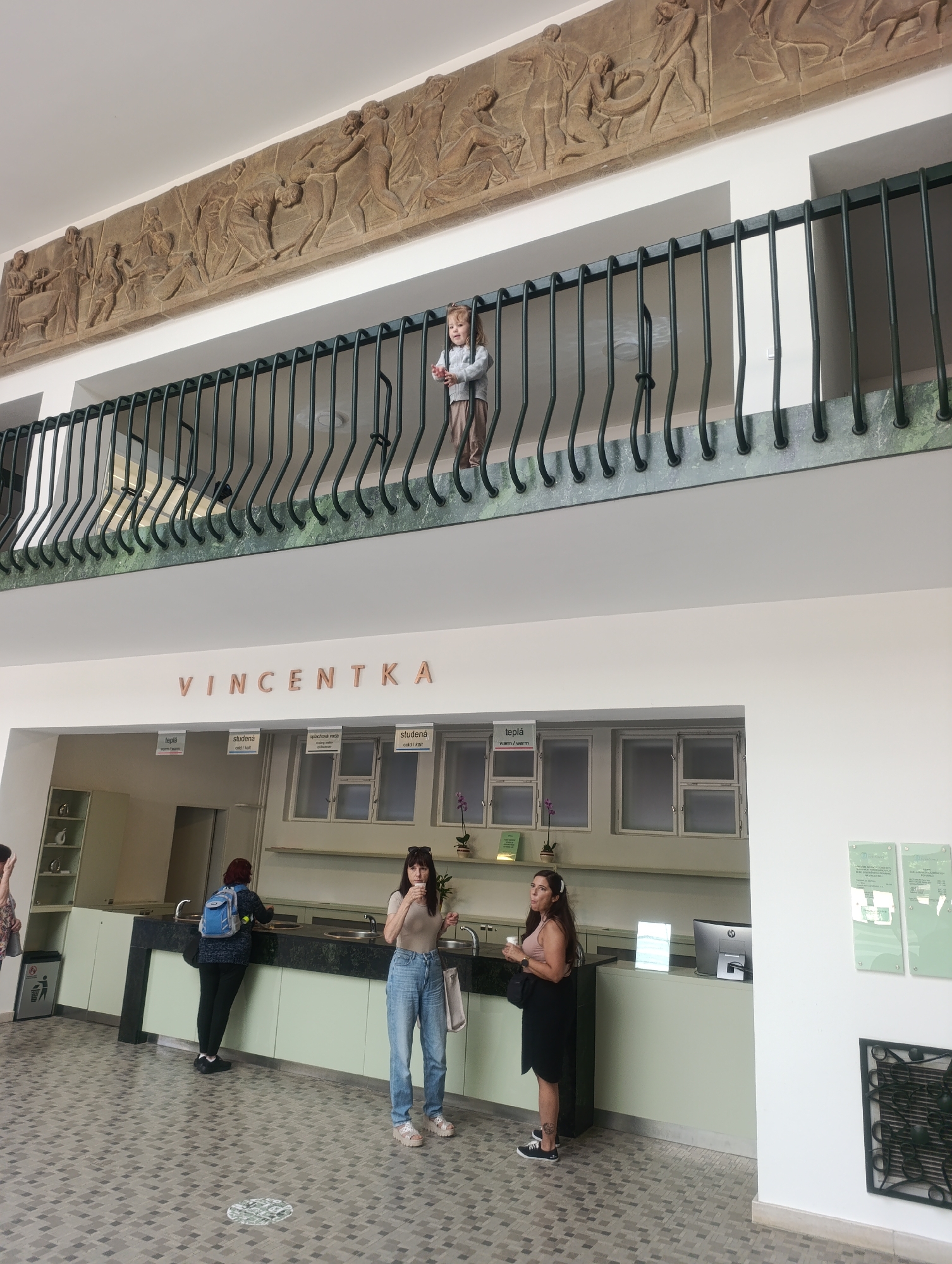 The image depicts an interior view of a building with a mezzanine level. On the mezzanine, a young child with blonde hair, wearing a light-colored sweater and pants, stands near a railing, looking downwards. Below the mezzanine, a long counter with sinks is visible, with three women present. One woman in the foreground on the right, wearing a light brown t-shirt and blue jeans, holds a cup to her lips. To her left, another woman, in a light brown short-sleeved top and dark skirt, also holds a cup. Further down the counter, a third woman, wearing a dark jacket and black pants with a blue backpack, is at a sink. Above the counter, the word "VINCENTKA" is prominently displayed in raised letters. The floor is tiled with a geometric pattern of grey squares and rectangles. The scene appears to be a public space, possibly a spa or a recreational facility, given the presence of drinking fountains and the name "VINCENTKA," which is known for its mineral springs in Luhačovice, Czechia. The walls are painted white, and the mezzanine railing is dark metal with decorative curved elements. A bas-relief frieze depicting figures is mounted on the wall above the mezzanine.
