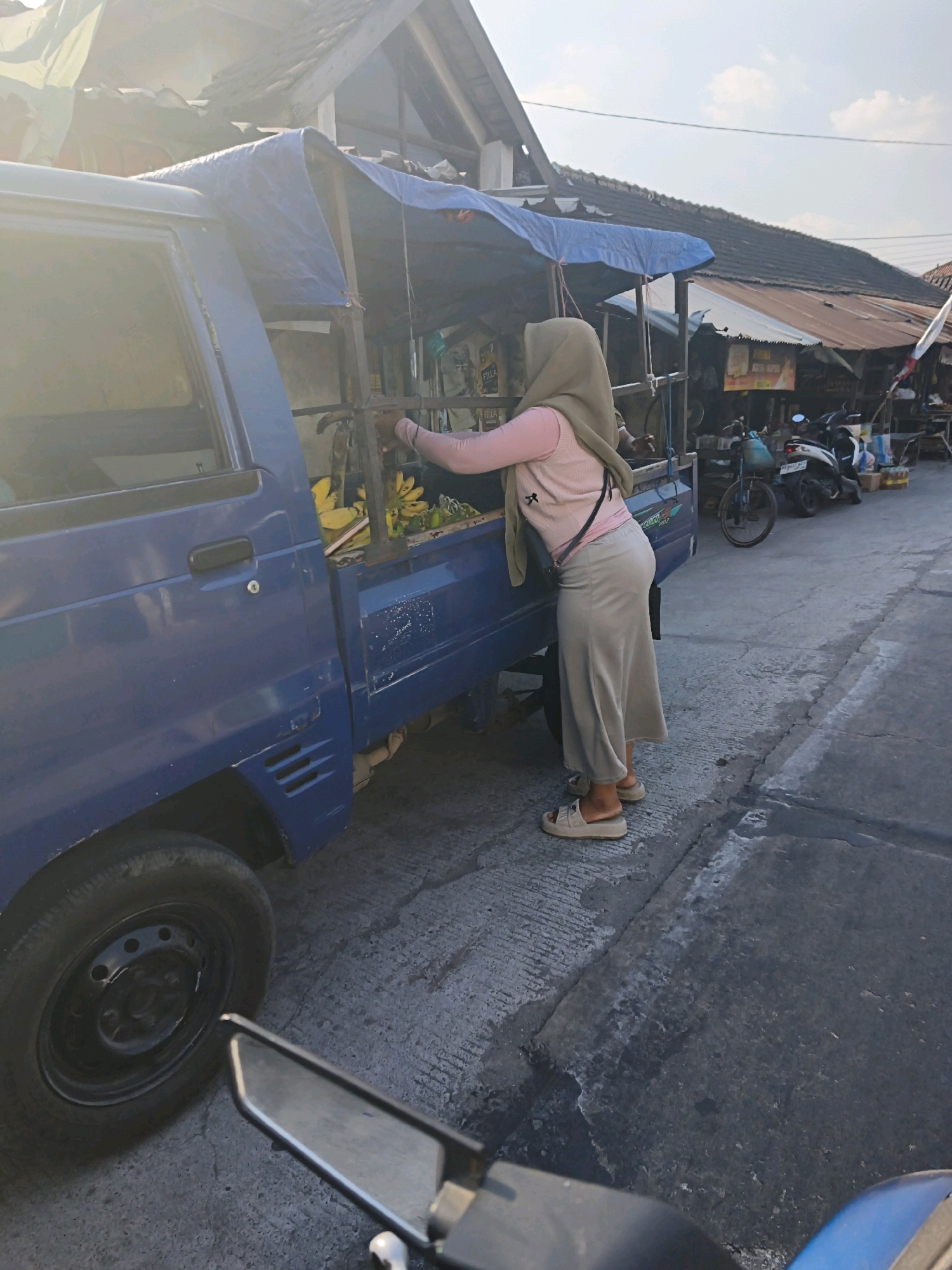 In Desa Dibal, Indonesia, a woman wearing a light green hijab and a pink long-sleeved shirt stands beside a blue pickup truck. She is wearing a long beige skirt and light-colored sandals. The woman is reaching into the back of the truck, which is covered by a blue tarp and appears to be carrying bananas. The truck is parked on a paved road in front of a row of buildings with corrugated metal roofs, suggestive of a market or commercial area. Several motorcycles are parked nearby, and the sky is visible with some clouds. The scene suggests a typical street market environment.