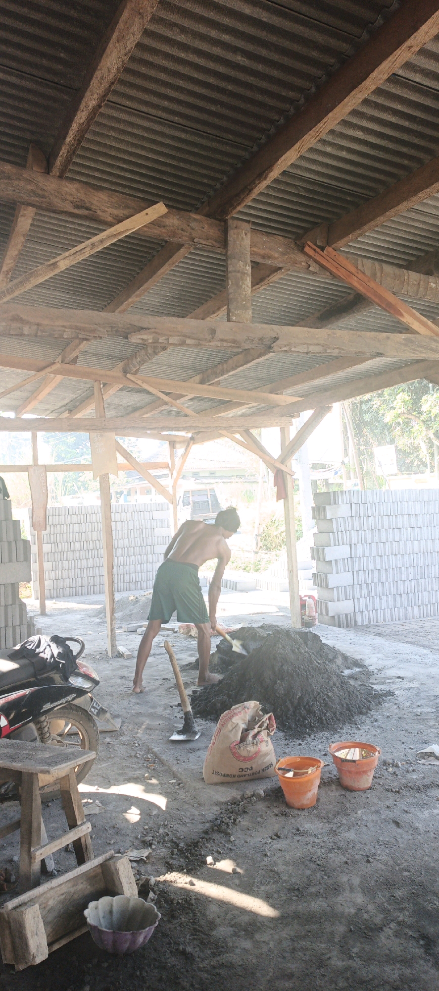 A man is working at a construction site in Kasiyan, Indonesia. He is wearing green shorts and is shirtless, bent over a pile of wet cement, using a shovel to mix it. The site is covered in dirt and has stacks of gray bricks on either side. A motorcycle is parked to the left, and a small wooden stool is beside it. In the foreground, there is a bag of cement, two orange buckets, and a purple bowl. The construction is under a roof made of corrugated metal supported by wooden beams. The sun is shining brightly, casting shadows across the scene.