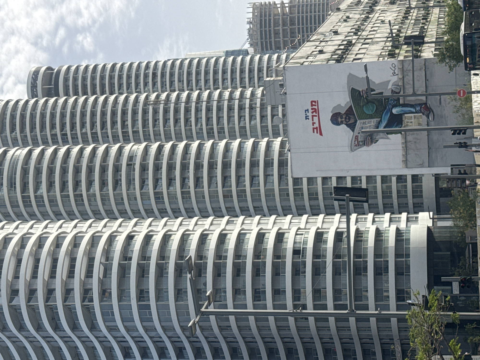 The image depicts a section of a skyline in Tel Aviv, Israel. It features two prominent architectural structures: a row of modern, multi-story buildings characterized by curved, wave-like balconies, and a taller building under construction with a raw concrete frame. A large billboard is attached to one of the completed buildings, displaying an advertisement with a man reading a newspaper. Traffic lights, street lamps, and sparse greenery are visible along the right edge of the frame. The sky is overcast with light clouds.