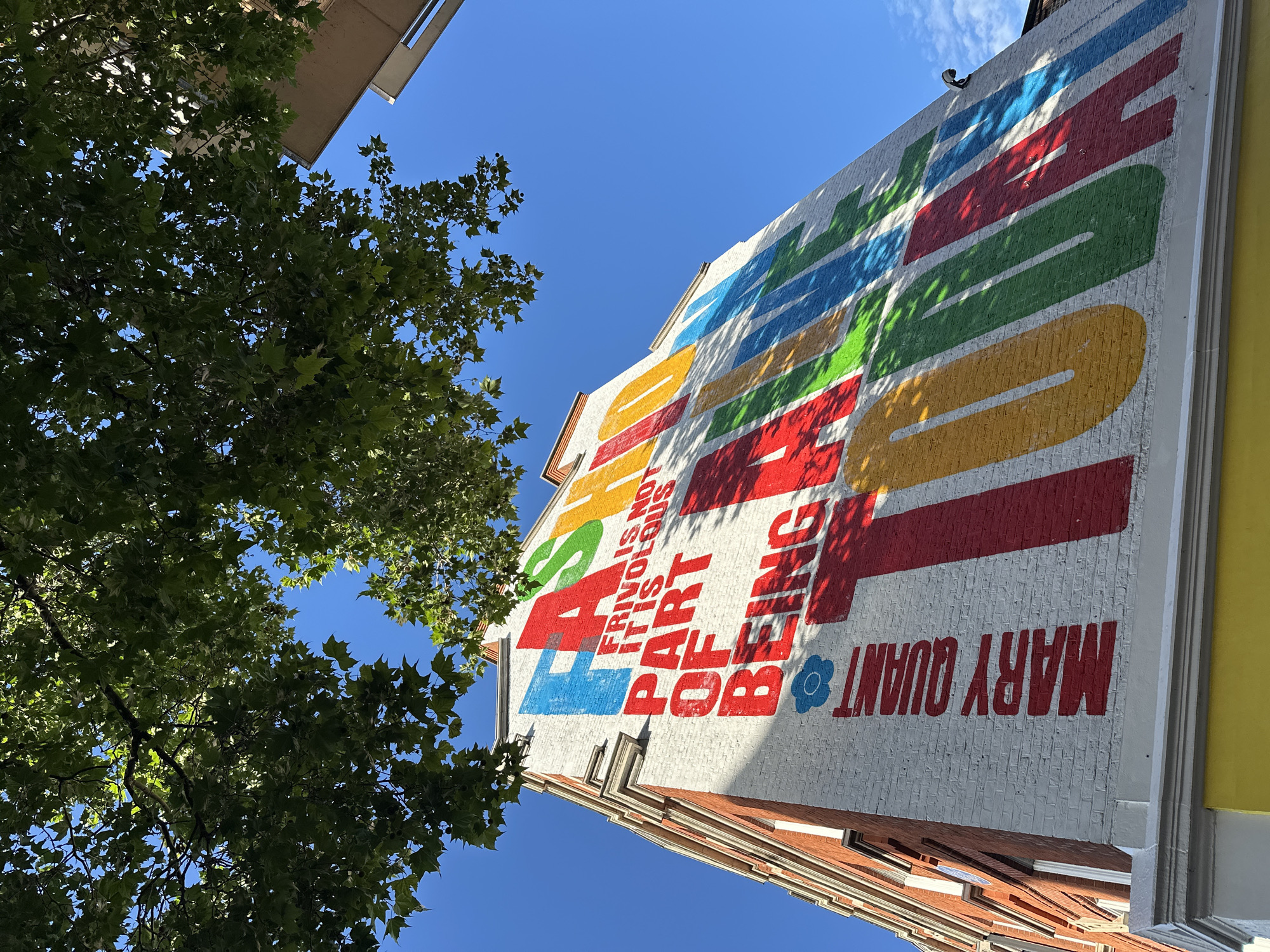 A low-angle shot captures a vibrant mural painted on the side of a white brick building in London, United Kingdom. The mural features large, colorful block letters that spell out "FASHION IS NOT FRIVOLOUS IT IS PART OF BEING ALIVE TODAY." The letters are painted in a spectrum of colors including red, orange, yellow, green, blue, and light blue, creating a bold and eye-catching display. The name "MARY QUANT" is written vertically in red on the left side of the mural, accompanied by a small blue flower symbol. Lush green tree branches with dappled sunlight are visible at the top of the frame, casting shadows on the mural and the building. The sky is a clear, bright blue with a few wispy clouds. The building itself has red brick accents and white architectural trim. Part of an adjacent building, a light brown rectangle, is visible on the right side of the image. The scene appears to be set on a sunny day.