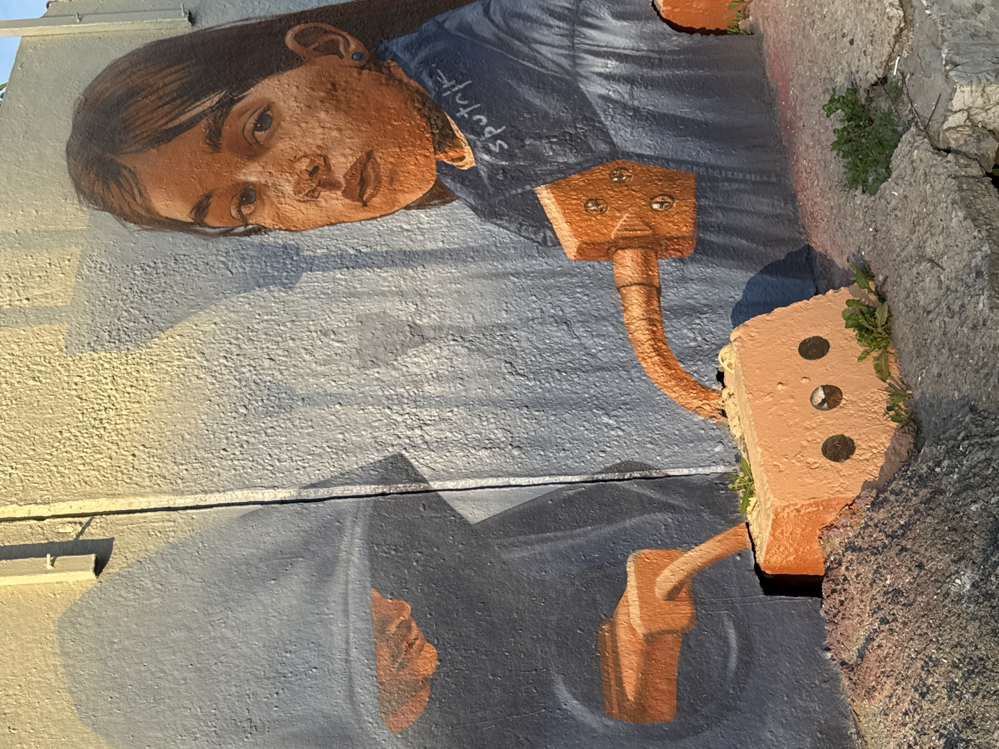 The image depicts a mural located in the city of Moscow, Russia, painted on a textured wall under direct sunlight. The wall's upper left section is illuminated with a warm yellow hue, transitioning to a light blue-grey texture across the rest of the surface. A distinct vertical seam or crack runs down the center of the wall.
The central figure is a young girl, appearing to be in her early to mid-teens, painted from the shoulders up, facing forward with a direct, somewhat pensive gaze. Her skin is rendered with an orange-brown, highly textured finish, mirroring the wall's surface. She has dark, straight hair parted on her right side, flowing over her shoulders. Her eyes are light blue, and she has a small blue stud earring in her visible right ear. She wears a dark blue shirt, on the right shoulder of which the artist's signature, "sputnk," is written in white script.
Several orange, brick-like, mechanical components are integrated into the mural. One such component extends from the girl's lower chest area, curving downwards and to the left. This piece features three visible metallic-looking rivets or bolts. It connects directly to a prominent orange brick object resting on the uneven, concrete ground at the bottom center of the frame. This ground-level brick has two dark circular depressions on its front face, resembling eyes. Small clusters of green weeds grow from cracks in the concrete surrounding this brick.
On the left side of the mural, a shadowy, dark blue-grey figure is depicted in profile, seemingly reaching down. From what appears to be its hand or lower arm, another orange brick-like connector extends horizontally, then curves downwards into a dark opening or crevice in the wall. A pile of dark, granular material, possibly dirt or gravel with reddish undertones, is present on the ground below this section. Another smaller, orange brick-like object is visible within the darker blue-grey area on the far left. At the top left, a light-colored, horizontal conduit or pipe is affixed to the wall, casting a shadow.