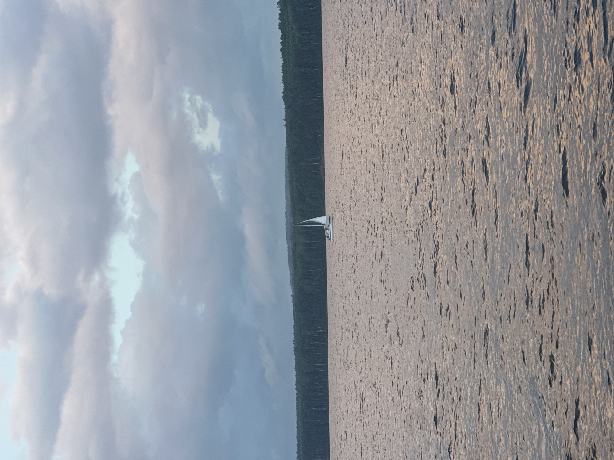 A sailboat with white sails is navigating across a body of water, likely a reservoir, as the sun begins to set. The water is a deep, shimmering hue, reflecting the soft, muted colors of the sky, which is dominated by a vast expanse of gray and pale blue clouds. The distant shoreline is lined with a dense, dark green forest, creating a stark contrast against the water and sky. The overall atmosphere is serene and tranquil, suggesting a quiet evening on the water. The scene is evocative of a peaceful moment in nature, away from the hustle and bustle of urban life. It is plausible that this scene is located near Chaikovsky, Russia, a city known for its water resources and surrounding natural beauty.