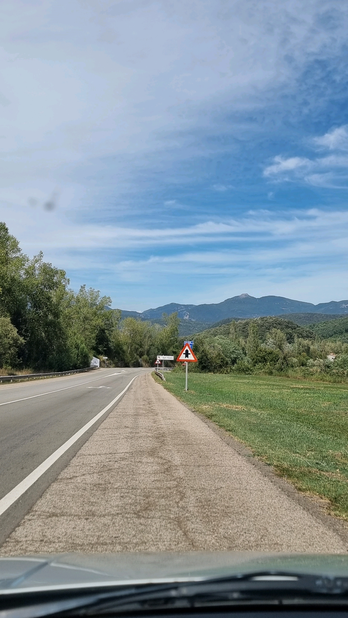This image depicts a road curving to the left, bordered by a paved shoulder and a grassy field on the right. Trees line the left side of the road, and a range of mountains forms the backdrop. A traffic sign indicating a merge ahead with a warning triangle is visible on the right side of the road. The sky is blue with scattered white clouds. The viewpoint appears to be from a vehicle. The location is identified as Sant Joan les Fonts, Spain.