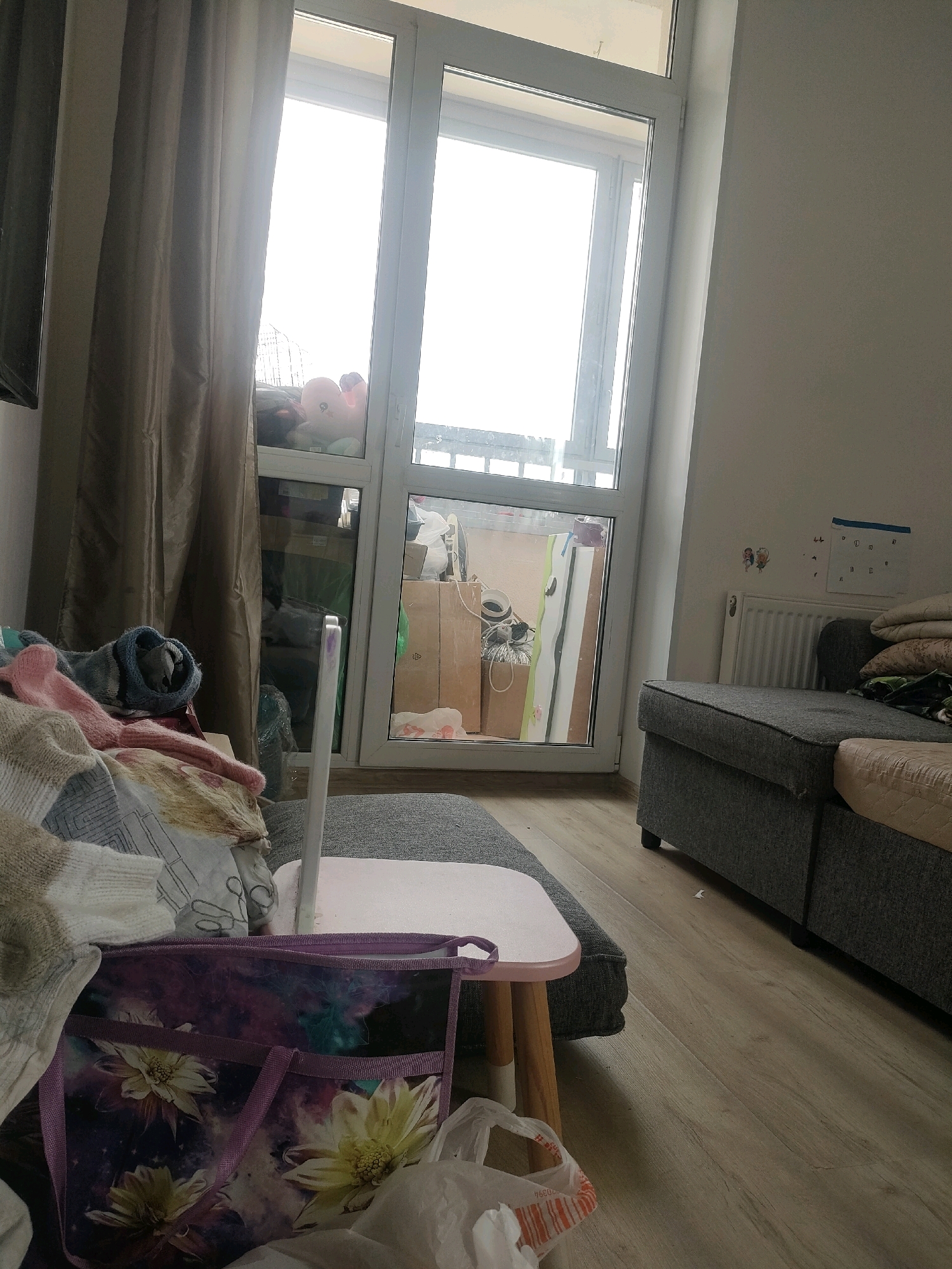 The image depicts an indoor scene, likely a living room or bedroom, with a large window or glass door leading to an outdoor balcony. The overall impression is one of a cluttered and lived-in space.
In the foreground, a collection of blankets and textiles is piled on what appears to be a sofa or chaise lounge. A small, pink-topped table with wooden legs is placed in front of this seating area. On the table, a white stylus or pen is visible. To the left of the table, a purple tote bag with a floral pattern is positioned, and next to it, a white plastic bag.
The large window or glass door dominates the middle ground, offering a view of a balcony. The balcony appears to be used for storage, with several cardboard boxes and other items visible. A pink stuffed toy is resting on the sofa inside, near the window.
The background shows a light-colored wall with a radiator and some items affixed to it, including what looks like a calendar. A dark gray sectional sofa is visible on the right side of the image. The flooring is a light-colored wood laminate.
The lighting suggests it is daytime, with bright light coming from the window. The scene does not provide any specific details to pinpoint its location within Yekaterinburg, Russia.