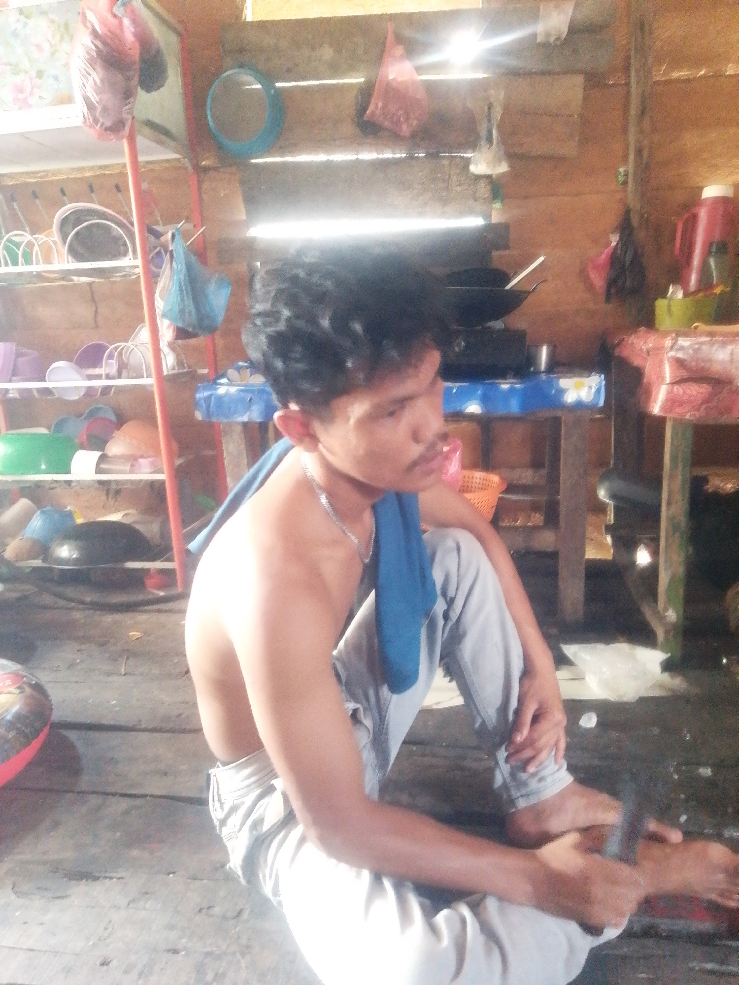 The image depicts an interior scene, likely a dwelling, with a young male sitting on a wooden floor. The man is shirtless, wearing light-colored pants, and has a blue cloth draped over his shoulder. He is positioned in the foreground, facing towards the right of the frame, with his legs crossed. His hands are visible, with one hand resting on his knee and the other near his ankle.

The background reveals a rustic environment with wooden walls and shelves filled with various items. On a shelf to the left, plastic containers and bowls are visible. Further back, a table is set up with what appears to be kitchenware, including a wok on a stove. A red container and a green bowl are also present on a table in the background. The overall impression is of a simple, functional living space. The lighting suggests it may be daytime, with light filtering in from an unseen source. The location is identified as Labuhan Bilik, Indonesia, a coastal town known for its historical significance and traditional way of life.
