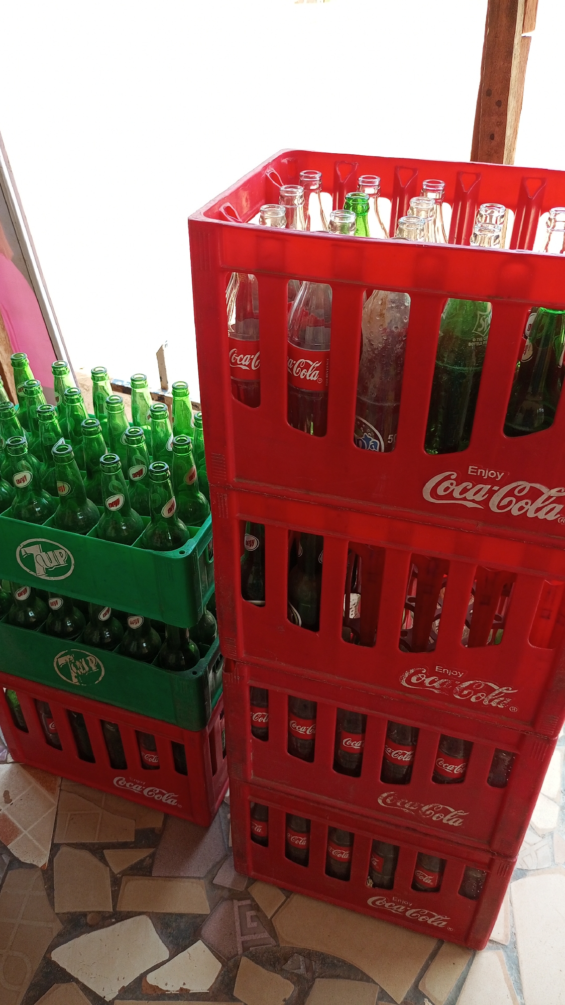The image shows a stack of four red plastic crates of Coca-Cola bottles next to a stack of two green and red plastic crates of 7-Up bottles. Some bottles are empty, while others still contain liquid. The stacks are located on a floor made of irregular, cracked tiles. The image was taken during the day, and the location is presumed to be Sabon Tasha, Nigeria.