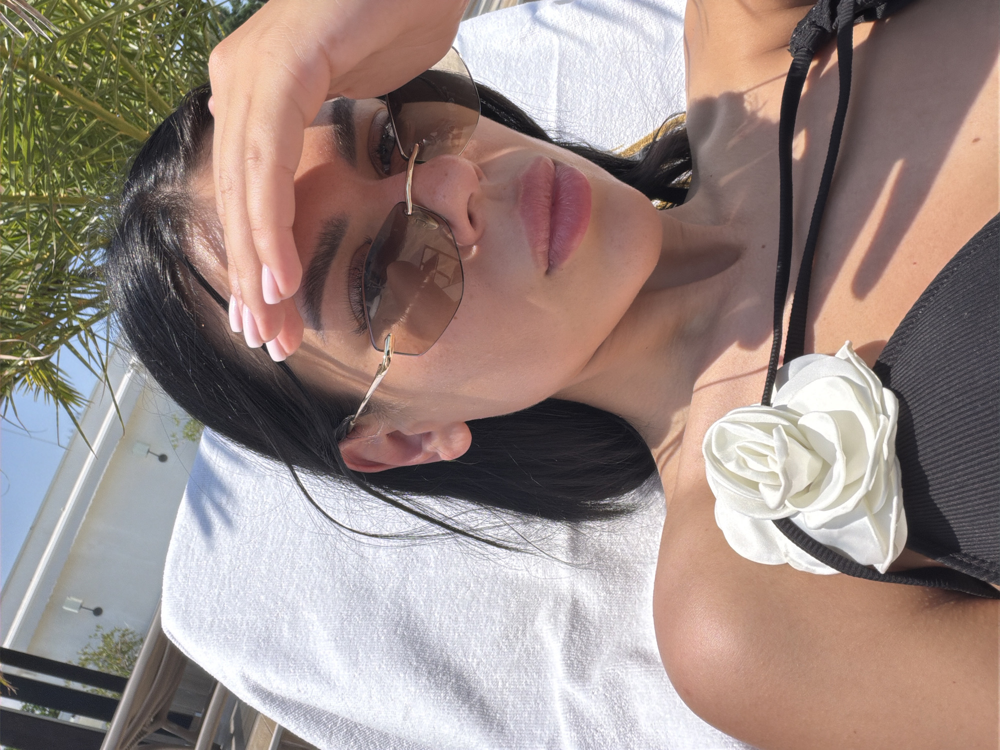 A close-up, high-angle photograph captures a woman relaxing outdoors, likely sunbathing, in Varna, Bulgaria. The subject is seen lying on her back on a white towel, which covers a sunbed or lounge chair. She has dark hair and wears brown-tinted sunglasses. One hand is raised, shielding her forehead and eyes from the bright sunlight, revealing light pink manicured fingernails. Her lips are full with a pink tint. She is dressed in a black bikini top, featuring a prominent white, camellia-like artificial flower attached to a strap near her chest.
The scene is illuminated by strong, direct sunlight, casting distinct shadows across her skin and the towel, indicating a clear, sunny day, possibly midday or early afternoon. In the background, lush green palm foliage is visible on the left side of the frame, suggesting a warm climate. To the left and higher up, a modern, white building with visible external light fixtures and architectural details stands against a clear blue sky. The overall impression is one of leisure and relaxation in an outdoor, resort-like setting. There is no visible text in the image.