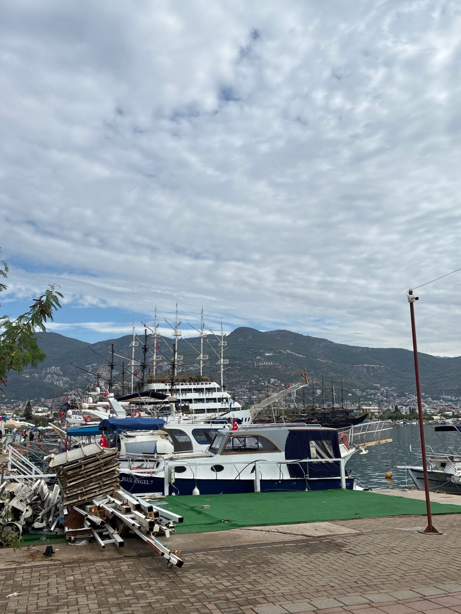 The image shows a view of Alanya Marina in Alanya, Turkey. In the foreground, there is a pile of scrap metal and wooden pallets on a brick walkway. To the right, a red pole with a security camera is visible. Several boats and yachts are docked in the marina, including a large, multi-masted sailing ship. The water is calm and reflects the cloudy sky. In the background, a city with white buildings is built on the slopes of a green mountain. A sign with the word "ALANYA" is visible on the mountainside. The sky is overcast with scattered clouds.