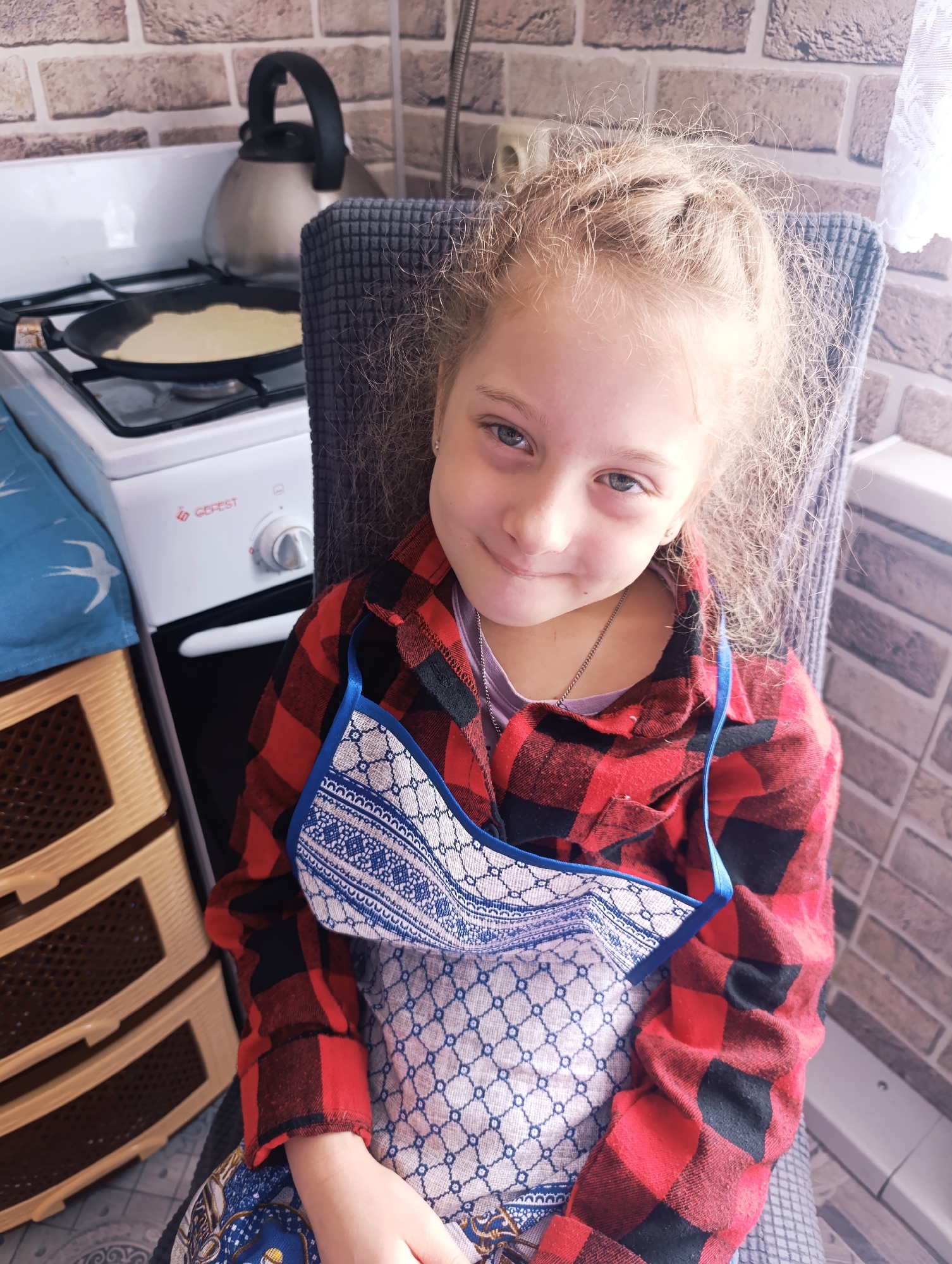 A fair-skinned young girl with blonde hair and blue eyes sits in a chair wearing a red and black flannel shirt over a purple t-shirt. She has a blue and white apron draped around her neck and resting on her lap. The girl is smiling slightly. Behind her is a grey chair. In the background to the left is a white stove with a black kettle on top and a frying pan with food cooking in it. Next to the stove is a beige plastic dresser. The wall behind the girl and stove is covered in a brick-patterned wallpaper. A white curtain hangs to the right. The location is a kitchen.