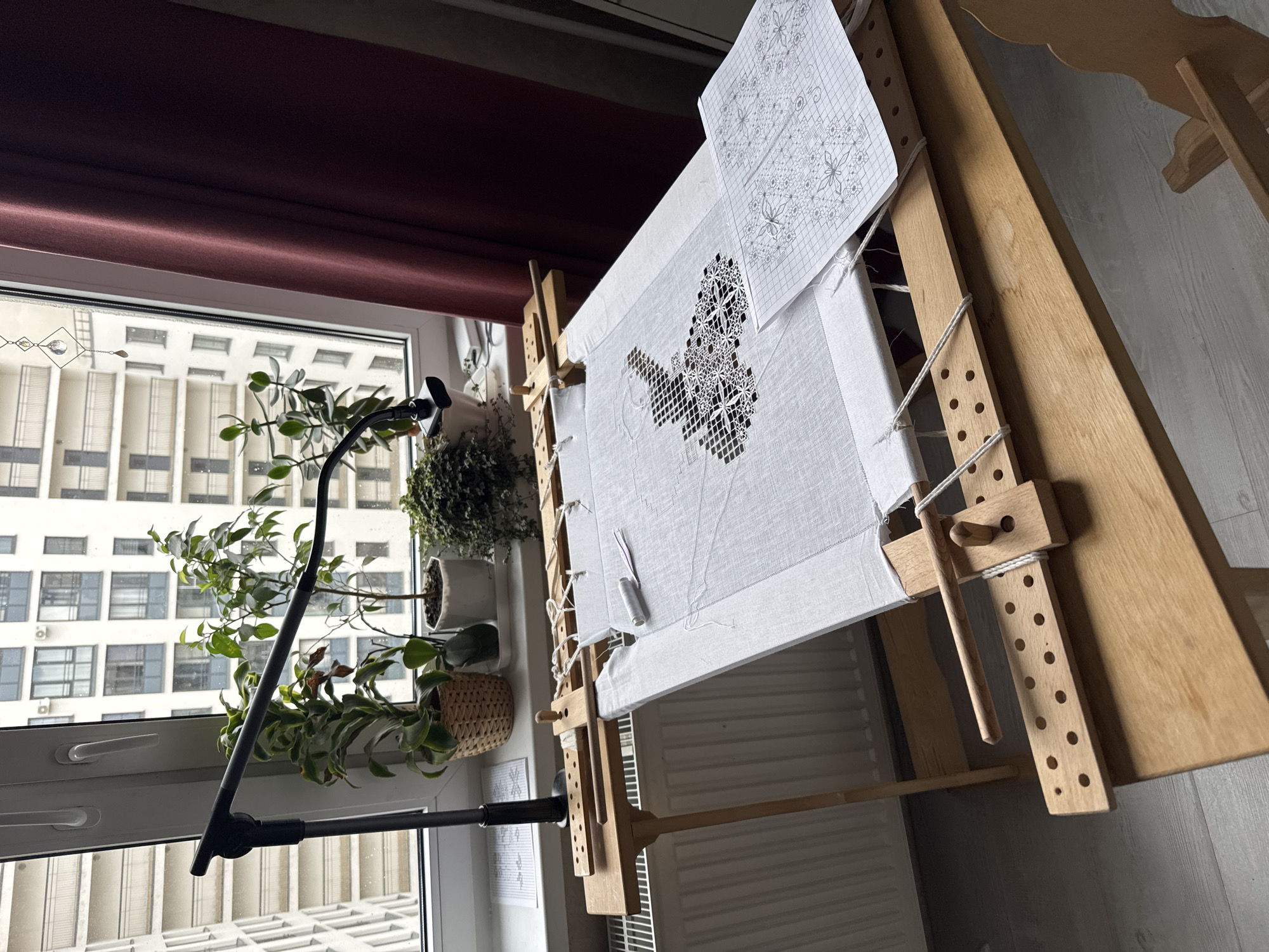 The image shows an embroidery project in progress, set up in a room near a window. The embroidery is stretched on a wooden frame, with a paper pattern pinned above it. The design appears to be a white-on-white geometric pattern. A lamp is positioned to illuminate the work area. Outside the window, a view of an apartment building is visible. The presence of plants on the windowsill adds a touch of nature to the scene. The building style seen through the window suggests the location is likely Saint Petersburg, Russia, considering the architectural style of the building. The room includes a radiator beneath the embroidery frame. The room is generally well-lit, suggesting daytime.