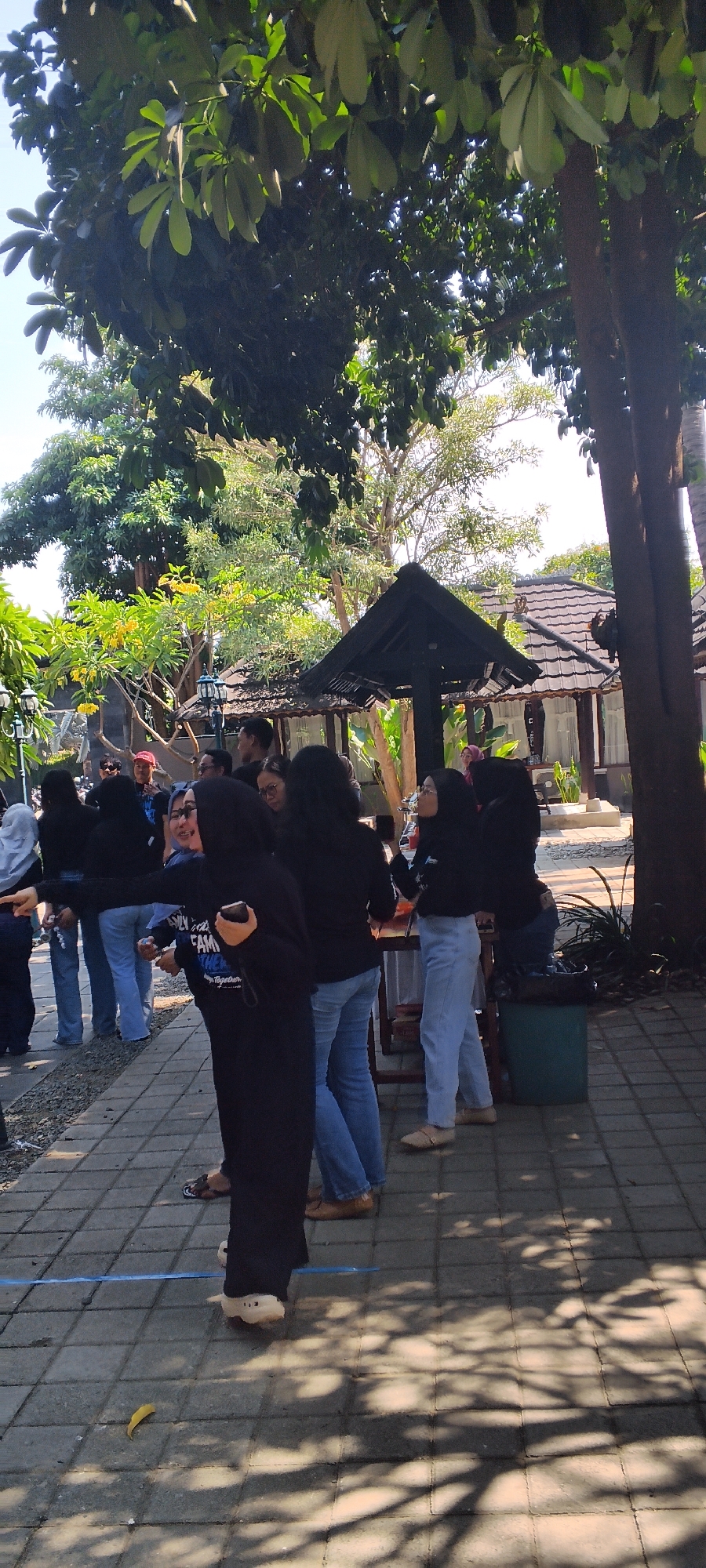 In Talise, Indonesia, a group of approximately ten to fifteen people, predominantly young adults, are gathered outdoors on a paved walkway lined with square gray bricks. They are dressed casually in dark tops, many appearing to be t-shirts or long-sleeved shirts, and various shades of denim jeans. Some individuals are wearing headscarves, indicating a mix of genders or cultural attire. The atmosphere seems relaxed as they stand and converse.

The setting is characterized by lush greenery. Tall trees with dense foliage cast dappled shadows across the walkway, suggesting a sunny day. In the background, traditional Indonesian-style buildings with dark tiled roofs are visible, hinting at a resort or a cultural complex. One building features a dark, pointed roof structure that resembles a gazebo or a covered pavilion. Scattered throughout the scene are elements of the environment, including a green trash bin and a small, fallen yellow leaf on the ground. The overall impression is of a casual outdoor gathering in a tropical or subtropical environment.