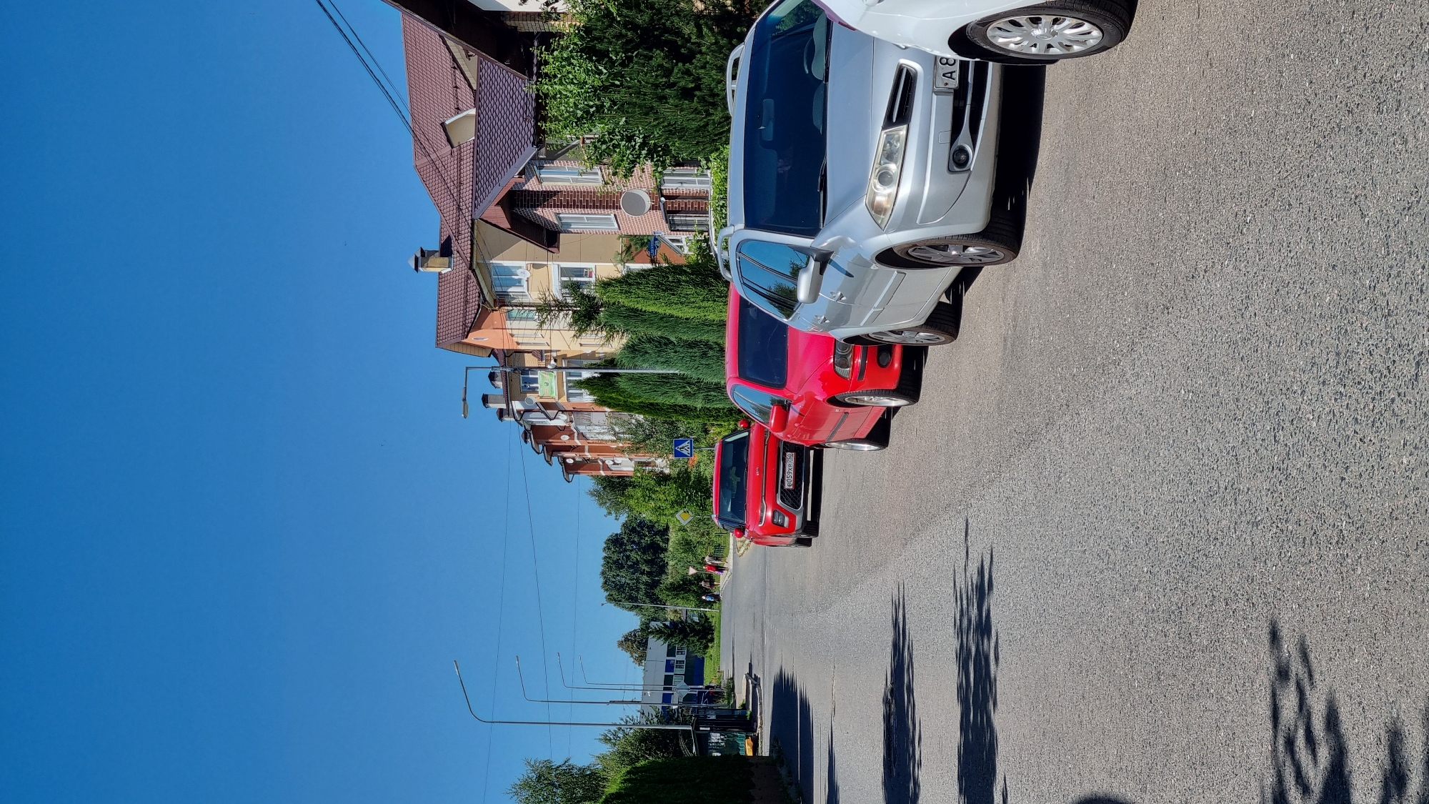 The image depicts a street scene in the city of Verzilovo, Russia, on a clear, sunny day. In the foreground, several cars are parked along the side of an asphalt road. The closest car is a silver station wagon, followed by a red sedan and a red SUV. The buildings lining the street are modern, multi-story residential structures with pitched roofs. Several evergreen trees are planted along the sidewalk in front of the buildings. In the background, the road continues, with more buildings and trees visible. There are a few pedestrians walking on the sidewalk in the distance. The sky is a vibrant blue.