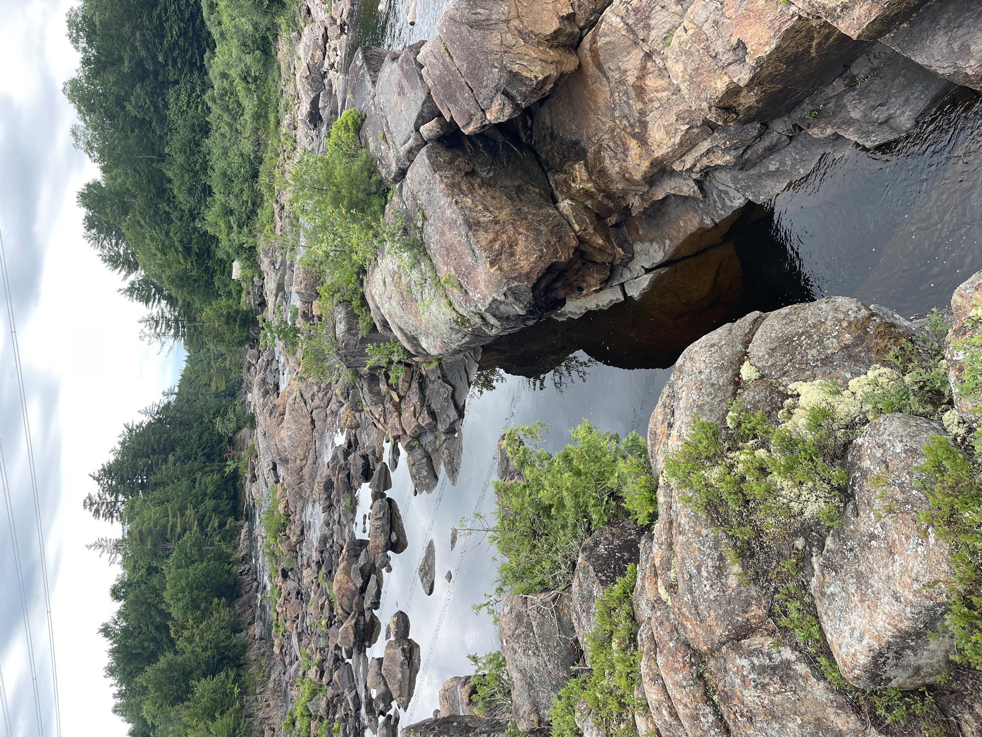 This is a vertical, outdoor, medium shot photograph taken in the Village of Lyons Falls, USA. The scene depicts a rocky riverbed with a dark, calm body of water flowing through it. Large, rough-hewn rocks, varying in size and color from reddish-brown to grey, dominate the foreground and midground. Green vegetation, including small trees and moss-like growth, is scattered amongst the rocks, with a dense forest of green trees lining the background to the left. The water's surface reflects the cloudy sky and the surrounding rocks. The overall impression is a natural, rugged landscape, possibly during the day given the lighting. There are no people or discernible objects other than natural elements and power lines visible in the far left of the image.
