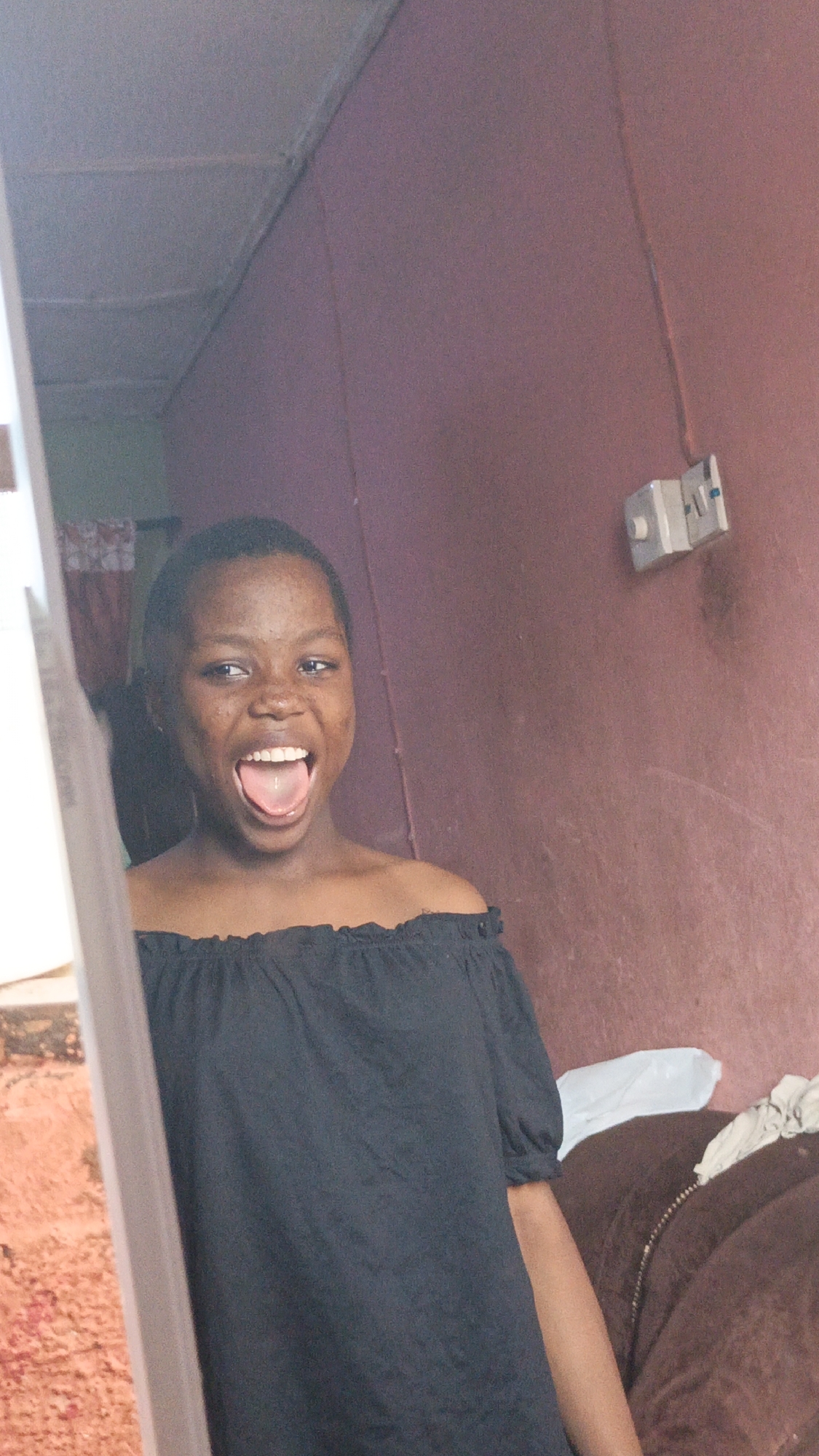 A young woman with short dark hair and brown skin is seen in a doorway. She is wearing a black off-the-shoulder top and is sticking her tongue out with a playful expression. Behind her, there is a purple wall with an electrical outlet. A brown couch with a white plastic bag on it is visible as well. The location is an interior, possibly a home in the city of Unknown, Unknown.