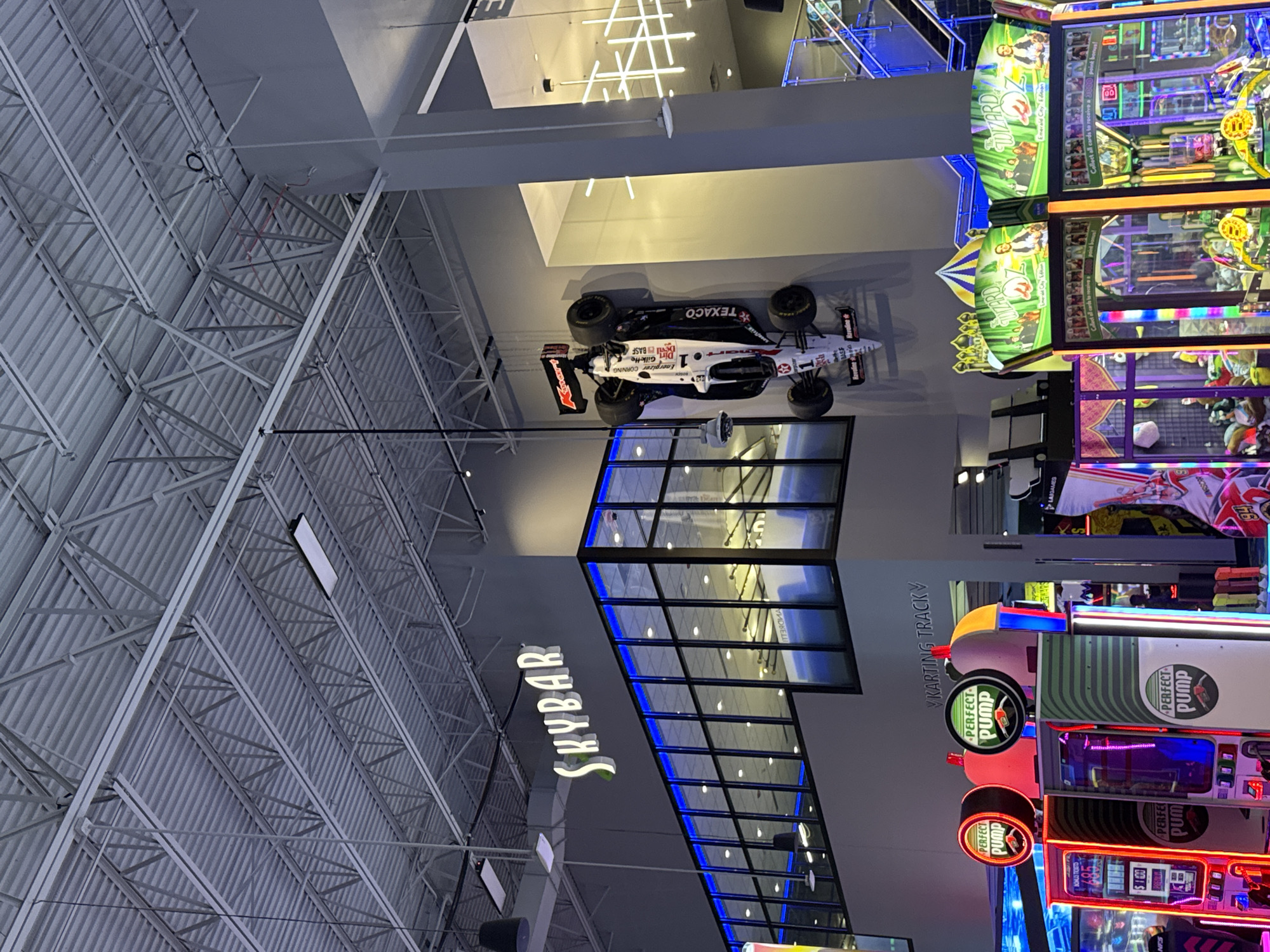This vertical, wide-angle shot captures a lively indoor entertainment complex in Schaumburg, United States of America. The high ceiling features an industrial design with exposed white metal trusses and corrugated panels.
A striking white race car, appearing to be a replica of an IndyCar or Formula 1 vehicle, is suspended from the ceiling. It prominently displays sponsor logos such as "TEXACO," "smart," "Energizer," and "Gillette," along with the number "1" and "GOODYEAR" on its tires. Other visible text on the car includes "CORNING" and "Pit Devil BASE."
To the left, a large, illuminated sign reads "SKYBAR," indicating a bar area. Below this, a modern architectural feature with square glass panes is highlighted by blue neon-like lighting. Further right, a sign for "VKARTING TRACKY" (likely "KARTING TRACK") suggests a go-karting facility.
The right side of the image is dominated by a vibrant array of arcade games. Several machines feature "PERFECT PUMP" branding with circular, illuminated signs. Another prominent game is "The WIZARD Emerald City Edition," with visible text encouraging players to "Collect all cards to receive a point bonus!" Other faint text seen includes "NFS," "LAIGAMES," and "NASDAQ." The bright lights and varied attractions indicate a bustling recreational environment. The indoor setting means the time of day is not discernible.