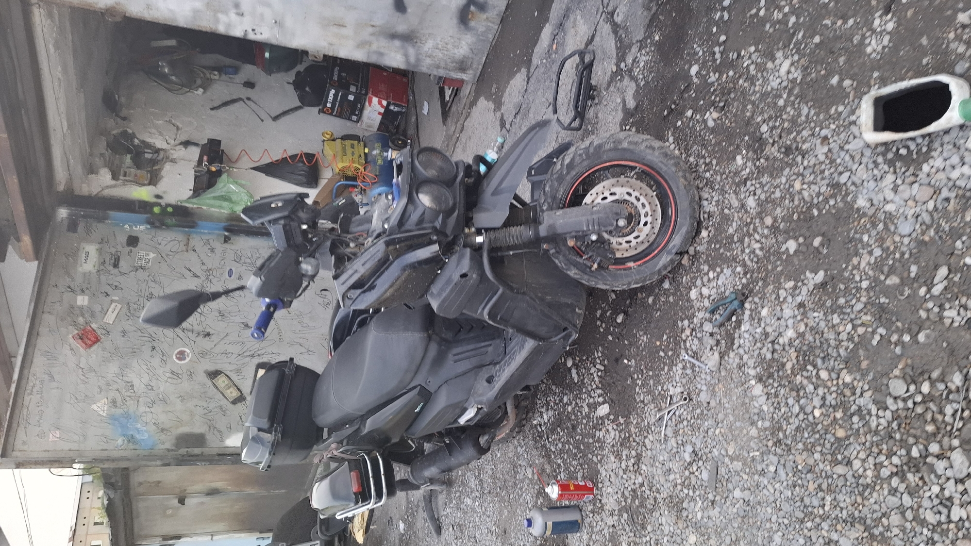 A dark-colored scooter lies on its side on a gravelly surface, possibly in a workshop or garage. The scooter is positioned horizontally in the frame, with its front wheel and handlebars to the left and the rear wheel to the right. The scooter has a large black seat, a black exhaust pipe, and black body panels. There are various tools and items scattered around the scooter, including a pair of pliers, a wrench, a can of spray, and a plastic container filled with dark liquid.
In the background, there is a wall covered in graffiti and stickers. Behind the wall, there are some boxes and electrical equipment, suggesting a workspace. The overall scene appears to be outdoors or in an open-air structure, given the gravel and open space. The lighting suggests it could be daytime. There is no indication of any people in the image. The presence of tools and the position of the scooter might imply that it is being worked on or has recently been involved in an incident.